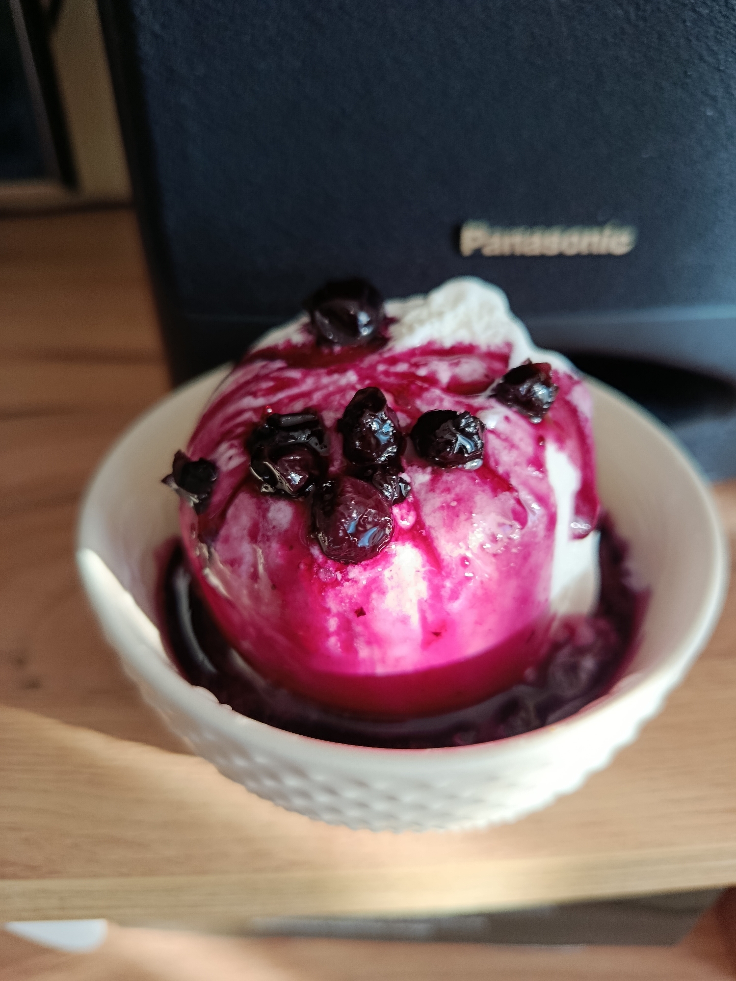 A close-up, eye-level shot shows a white, textured bowl filled with two scoops of ice cream, topped with a vibrant pink sauce and dark berries. The ice cream appears to be melting slightly, with the sauce cascading down the sides. In the blurred background, a dark object with the "Panasonic" logo is visible. The bowl rests on a light-colored wooden surface, with sunlight casting shadows across it. The scene is indoors, likely in a kitchen or dining area. There are no people or explicit actions depicted, and the image lacks any specific indicators of its location within the city of Minsk, Belarus.