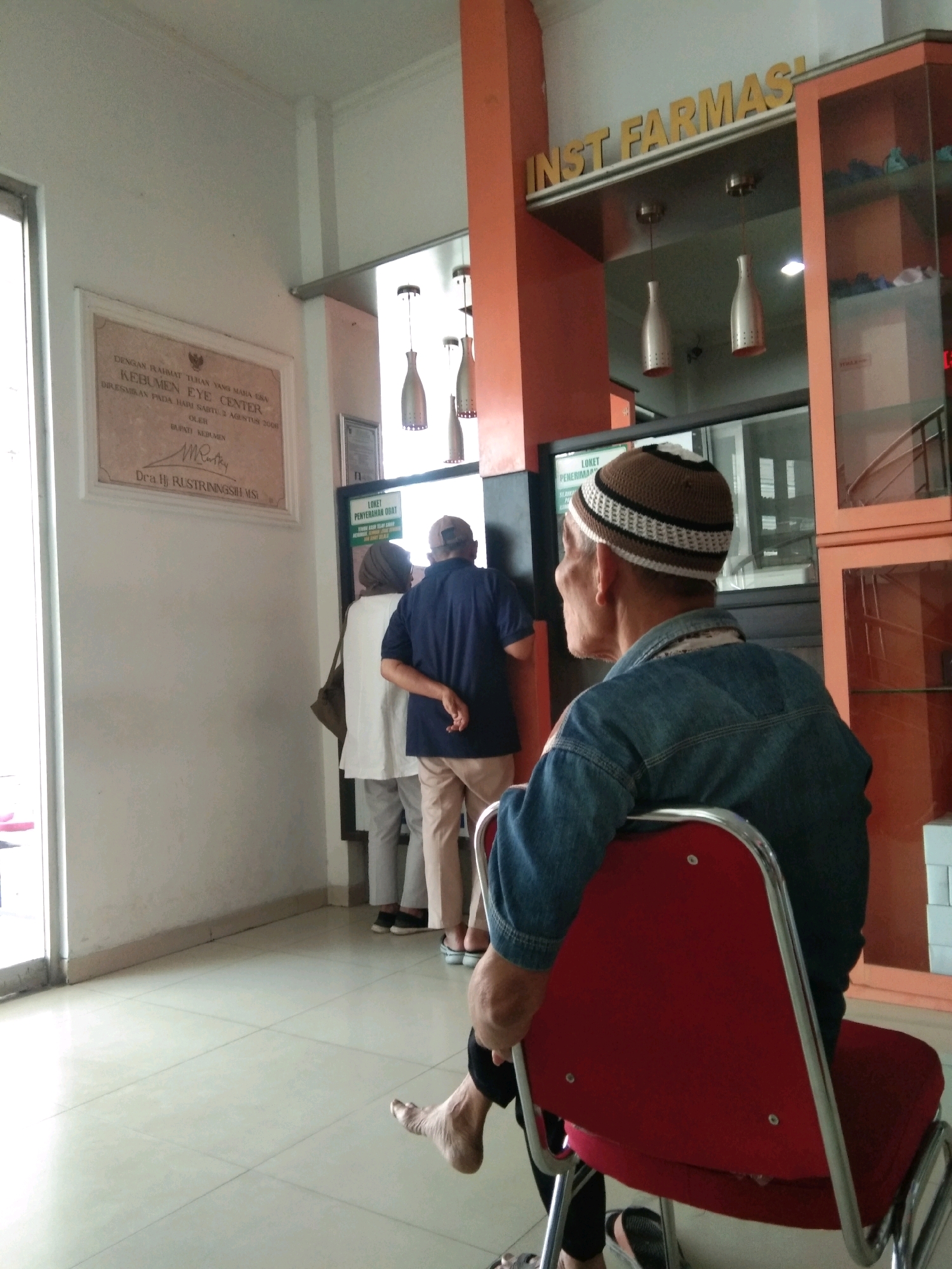 In the waiting area of the Kebumen Eye Center in Kebumen, Indonesia, several individuals are visible. A man in a blue denim jacket and a brown and white kufi hat is seated in a red chair, facing away from the camera. In the background, two individuals stand near a window, one wearing a white top and headscarf, and the other a dark blue shirt and light-colored pants. A sign reading "INST FARMAS" is prominently displayed above the counter. The room has tiled floors and white walls, with an orange architectural element to the right of the entrance. A framed document is mounted on the left wall.