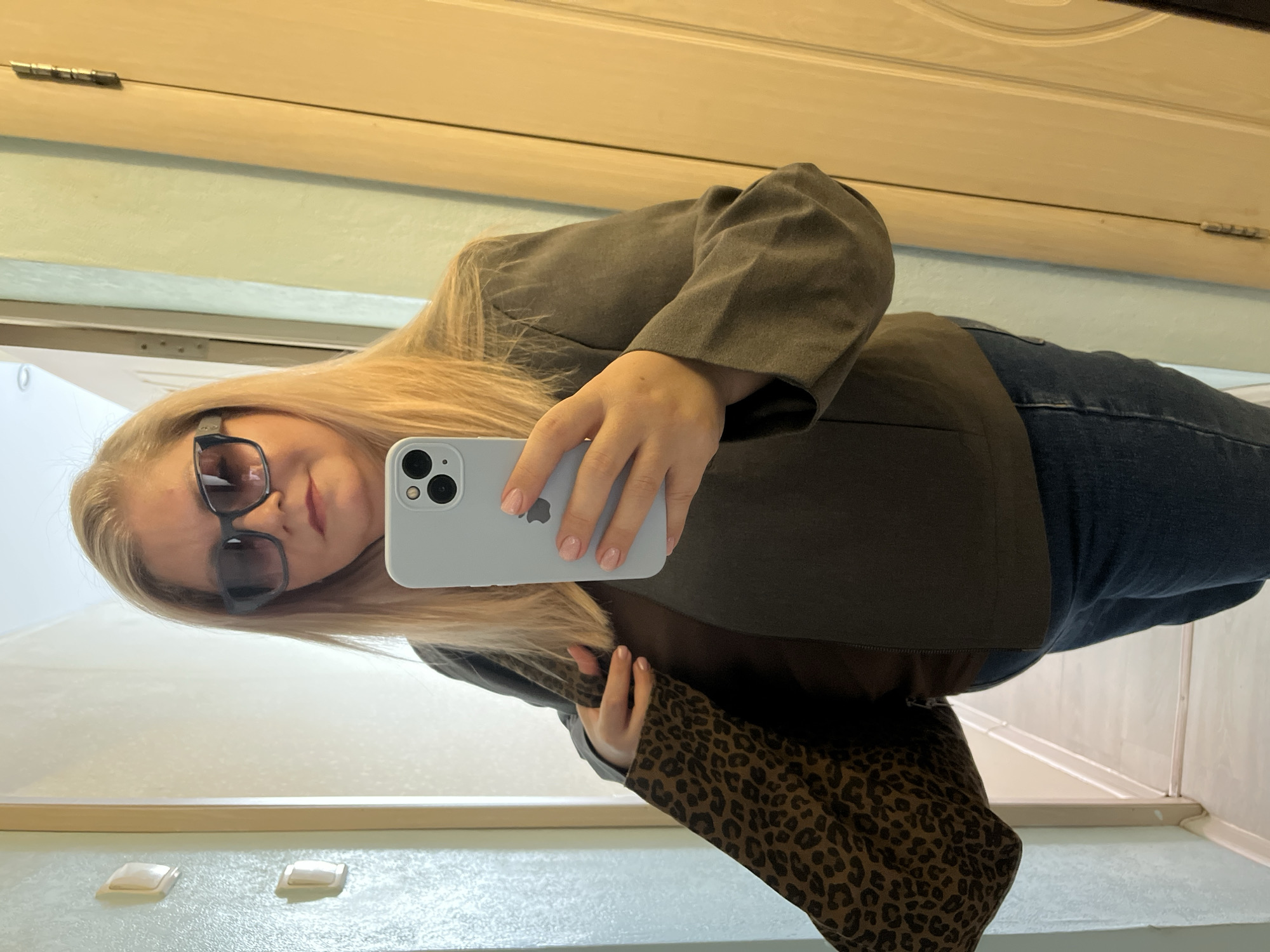 A woman with blonde hair and sunglasses is taking a selfie in what appears to be a bathroom. She is wearing a green blazer, a leopard print top, and jeans. She is holding a light blue iPhone to take the photo. The bathroom has a light green wall and a wooden door. There are light switches visible on the wall. It is not possible to determine the specific location within Izhevsk, Russia from the image.