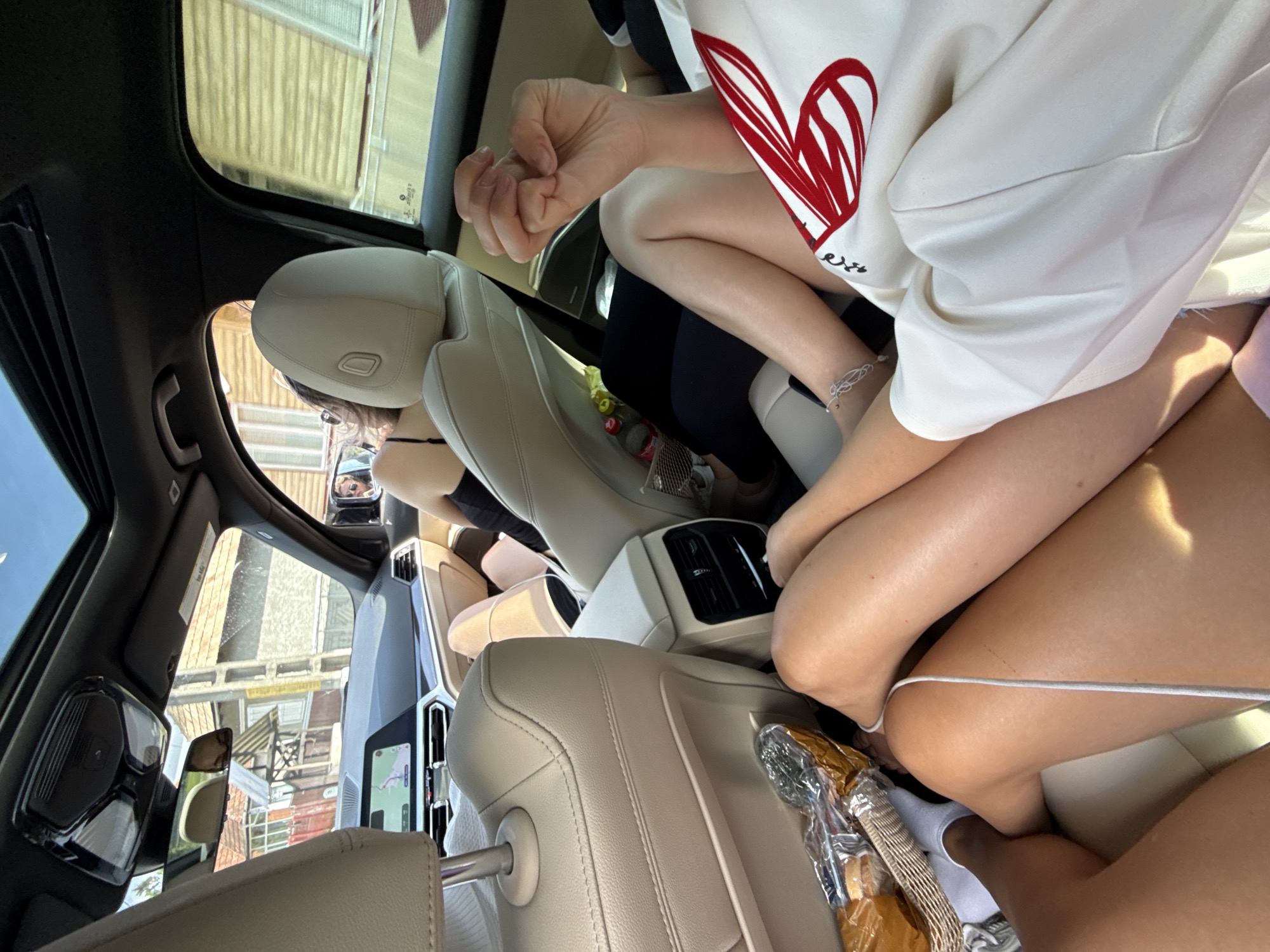 The image, taken in a car likely in the city of Hârja, Romania, shows the interior from a low-angle perspective, focusing on the legs of at least two individuals. The car has light-colored leather seats, a sunroof, and a digital display in the front. Outside the car, buildings with red-tiled roofs and wooden facades are visible, consistent with architecture found in Romania.
In the foreground, two pairs of legs are prominent. One set of legs is tanned, wearing white socks and positioned near a wicker basket containing bread. The other set of legs is also tanned and bare, crossed over the first. Another person's legs, wearing dark pants, are visible further back. A person with dark hair is seated in the passenger seat, wearing a black top.
A hand with clasped fingers is visible in the mid-ground, resting on the legs of the person wearing the white top with a large red heart design. The environment outside suggests a sunny day in a residential area. The overall scene depicts a casual setting inside a vehicle, possibly during a road trip.