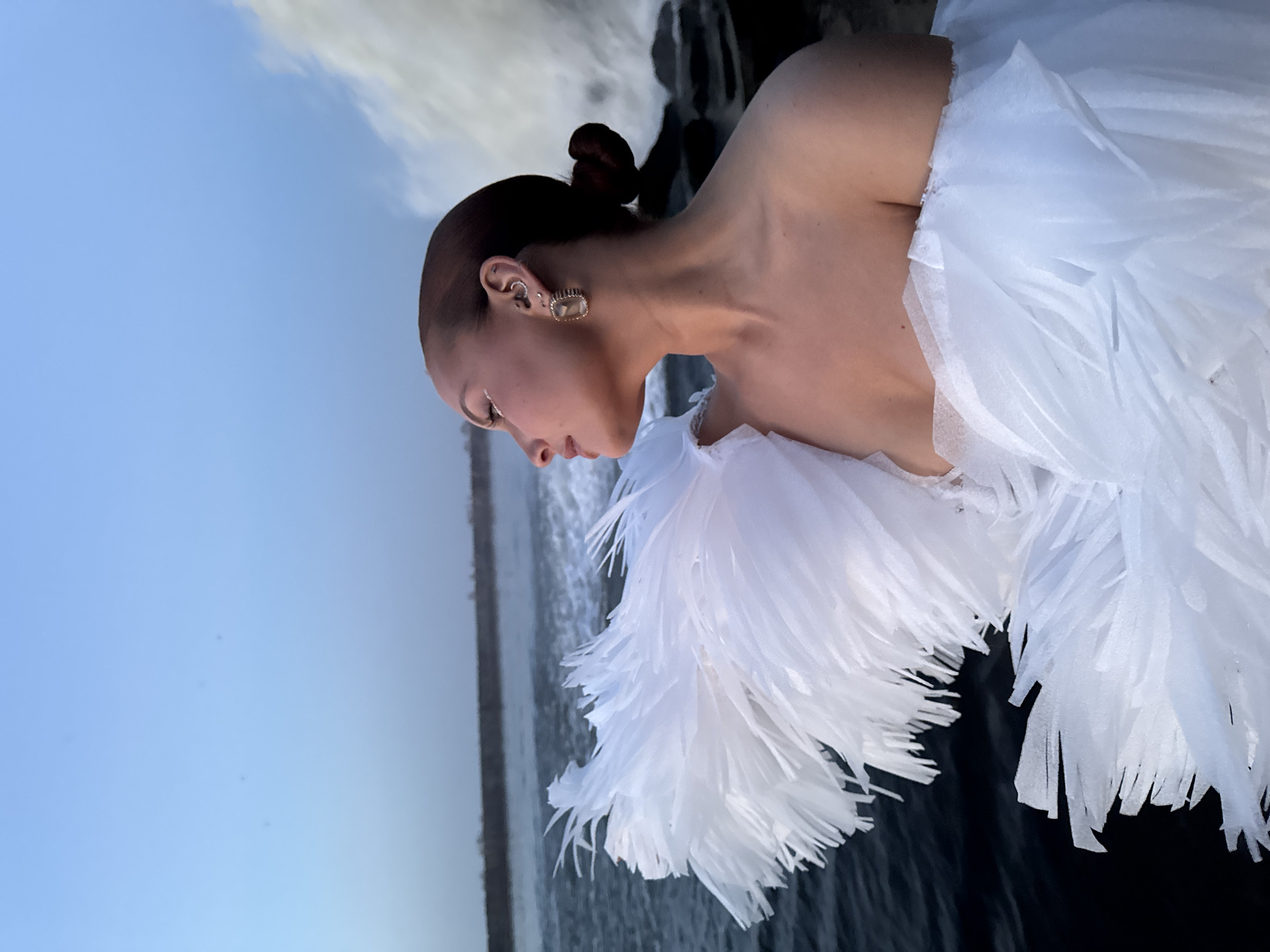 A sideways medium shot depicts a person with fair skin and dark hair pulled into a bun. They wear a white garment with many pointed frills. The person is gazing downward to the left. They wear earrings on both ears, one large and square. The backdrop is blue and black, indicative of sky and water. The city of Noviy, Russia is not discernible from the image.