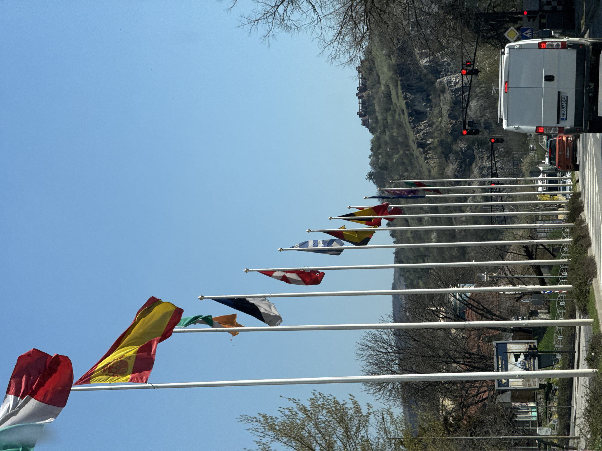 The image shows a street in Veliko Tarnovo, Bulgaria. A row of flagpoles displays various national flags. A white van with the license plate "EB 6973 BP" is stopped at a traffic light, which is red. A car is behind the van. The street is lined with trees, and a rocky hillside is visible in the background. There is a billboard and a sidewalk visible.