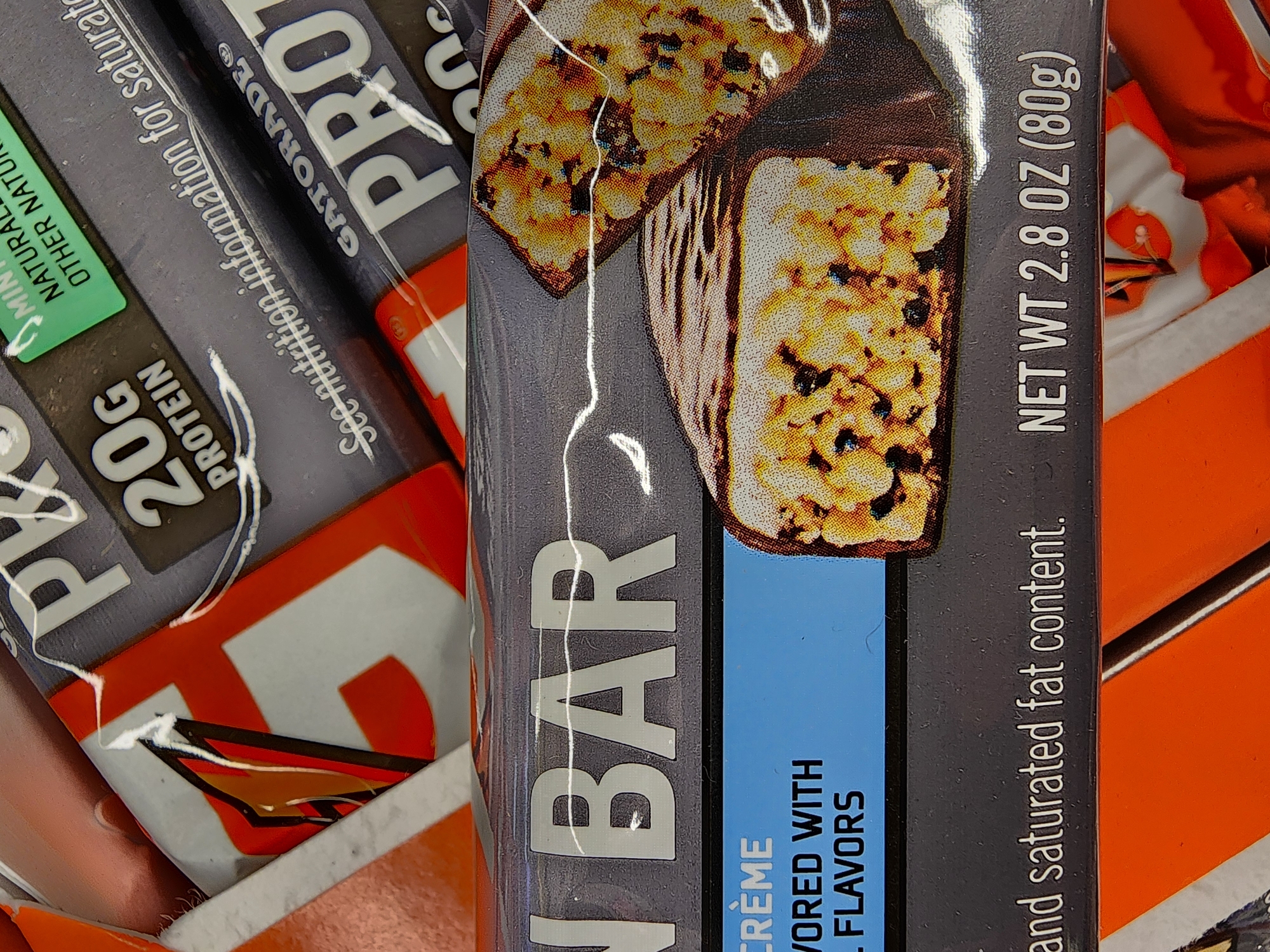 The image shows a close-up of several protein bars, with the focus on the packaging of a "PROTEIN BAR" and a "GATORADE PROTEIN" bar. The "PROTEIN BAR" packaging is dark gray with white text and features an image of two protein bars. Text on this packaging includes "CRÈME," "VORED WITH," and "L FLAVORS." Below this, it reads "and saturated fat content. NET WT 2.8 OZ (80g)." The "GATORADE PROTEIN" bar packaging is also visible, with "GATORADE" in large white letters and "PROTEIN" below it in even larger letters. There is also text on this packaging that says "See nutrition information for saturat." and a green label with "MIN NATURAL OTHER NATU." An orange and white graphic is visible on the left side of the image, partially obscured. The background appears to be a retail environment, possibly a grocery store shelf, with other packaging visible. The overall lighting is artificial and highlights the product packaging. There are no people or animals in the image. The setting is a retail environment, likely a store selling nutritional supplements or snacks. The image does not provide specific details that would allow for pinpointing a location within Camp Hill, United States of America, but it depicts consumer products in a commercial setting.