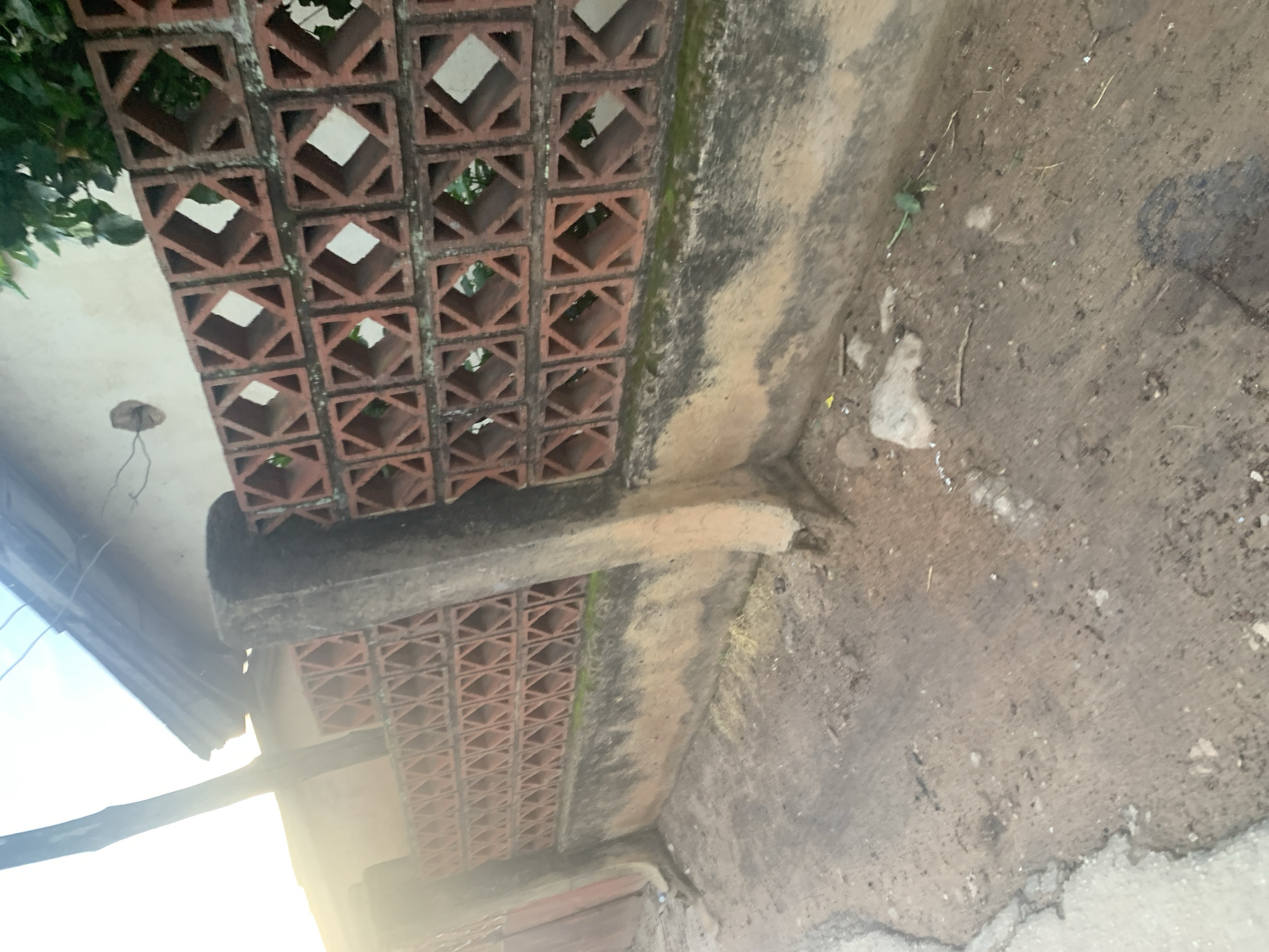 The image captures a sunlit outdoor scene, likely in the city of Jos, Nigeria, showing a textured, weathered wall constructed from reddish-brown bricks with a distinctive diamond-shaped cutout pattern. This wall runs horizontally across the frame and is supported by two rough, grey concrete pillars. The ground in front of the wall is a mix of dirt and debris, with some patches of light-colored gravel or broken concrete. To the left of the wall, a hint of a red gate or door is visible. Above the wall, the roofline of a building and some greenery can be seen, suggesting a residential or commercial area. The overall impression is one of an older, utilitarian construction, common in many parts of Jos.