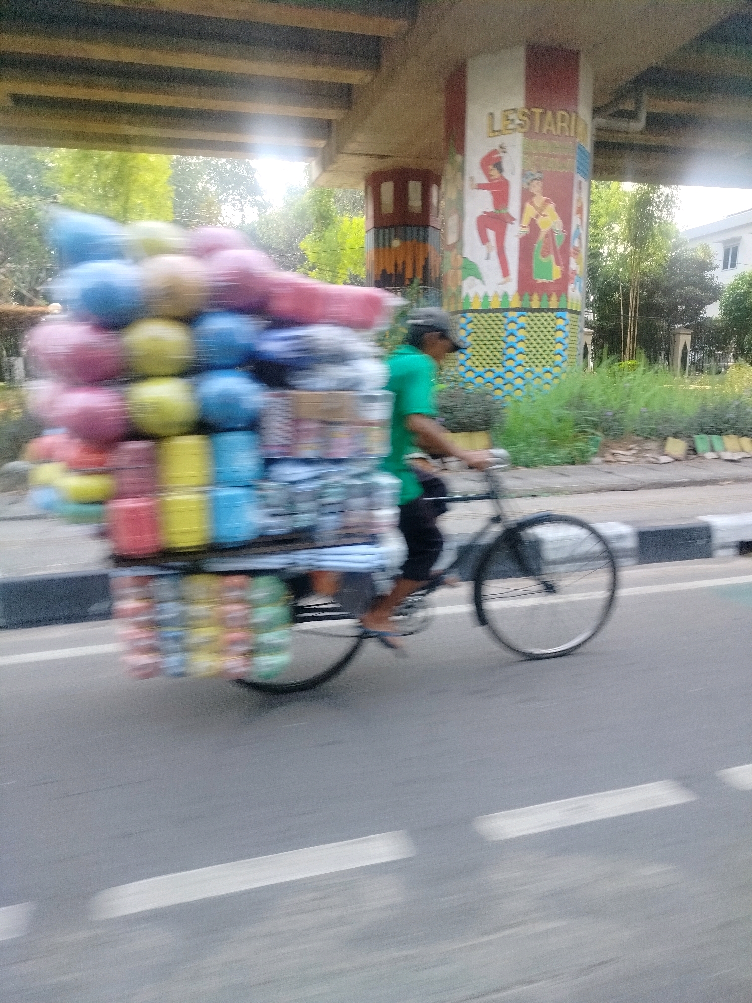 The image shows a man riding a bicycle loaded with merchandise under an elevated structure in East Jakarta, Indonesia. The man is wearing a green shirt and a dark cap, and is riding a bicycle with a large, multi-tiered cargo carrier laden with colorful plastic containers, possibly for sale. The elevated structure appears to be a road overpass, with a painted pillar featuring the word "LESTARI" and decorative murals depicting figures. The environment includes greenery and other vegetation along the roadside, and the scene is captured with motion blur, indicating movement.