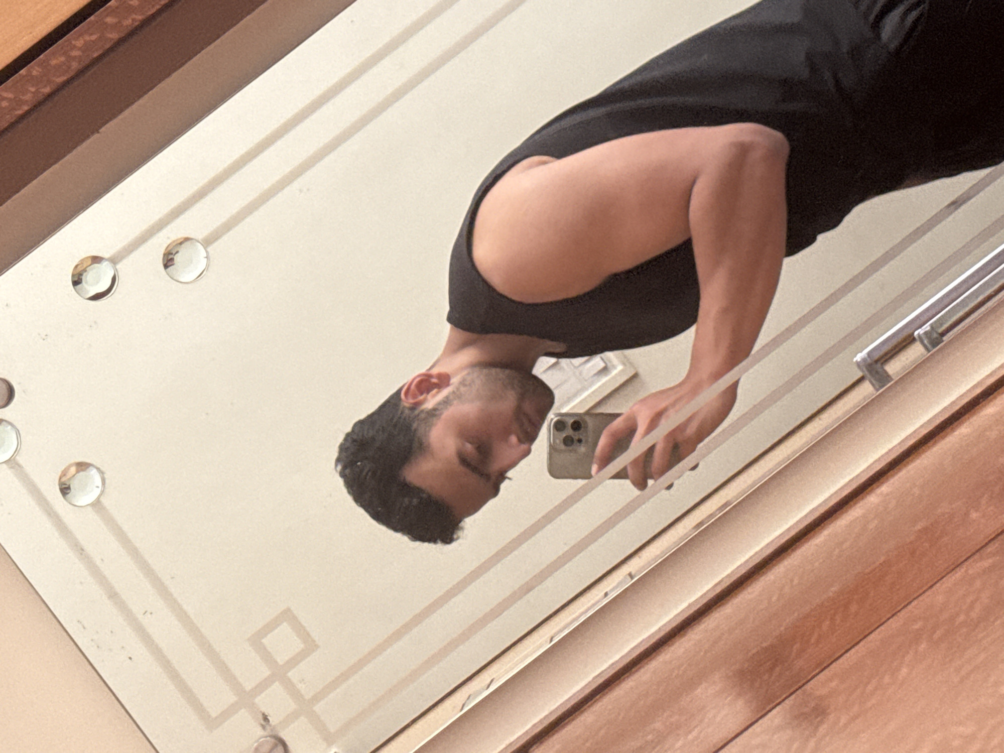A man with dark hair and a black tank top is taking a selfie in a mirror. The mirror has a white frame with decorative clear studs. The background includes wooden panels. Location: Unlikely to be Kolkata, India, based on typical architectural and interior design.