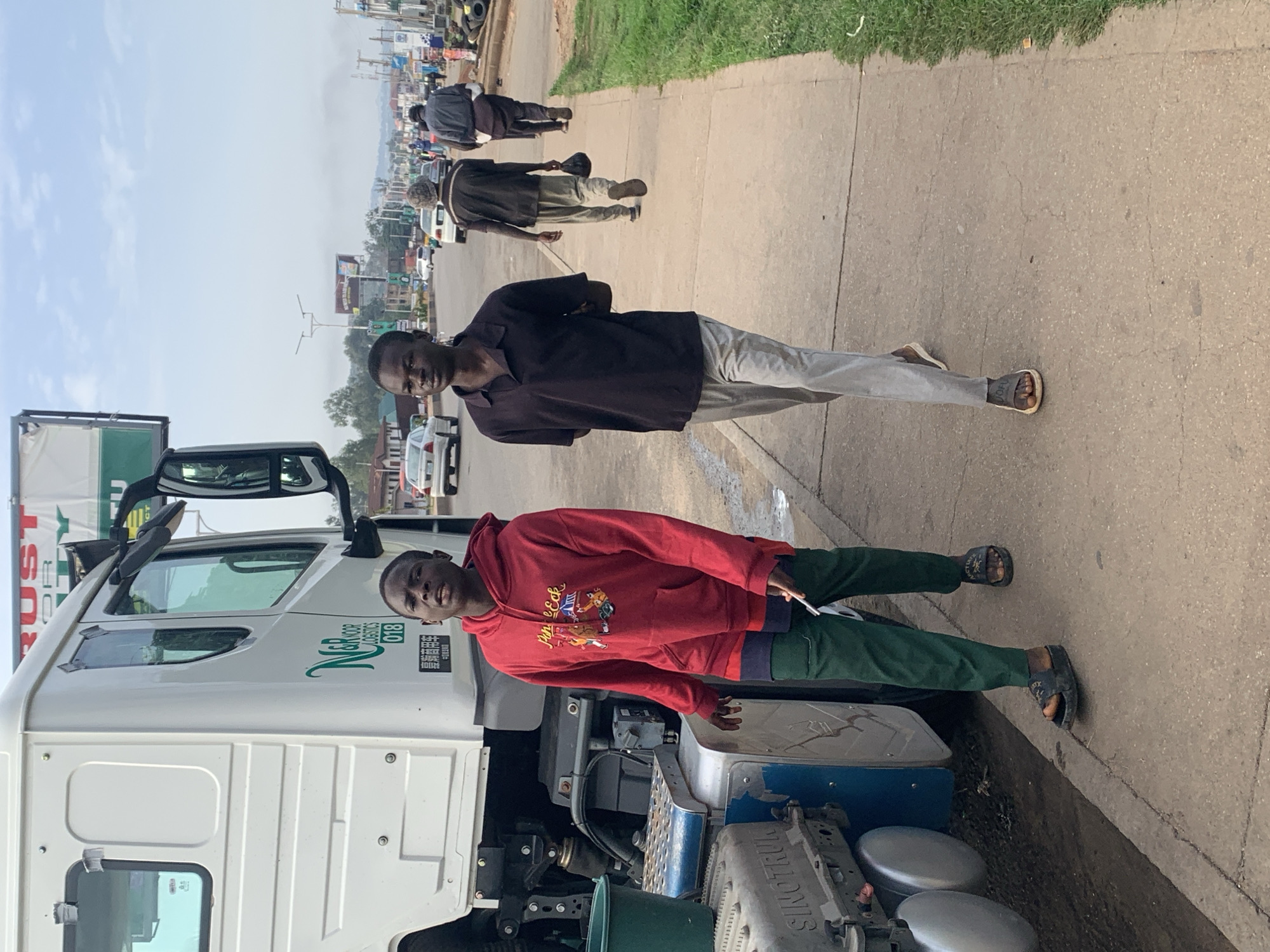 Two young men are walking on a sidewalk next to a parked white truck. The man on the left is wearing a red hoodie and dark green pants. The man on the right is wearing a dark polo shirt and light gray pants. The truck has "N&R LOGISTICS 018" written on its side. In the background, there are cars on a road, buildings, and streetlights. The scene appears to be in an urban or semi-urban area.