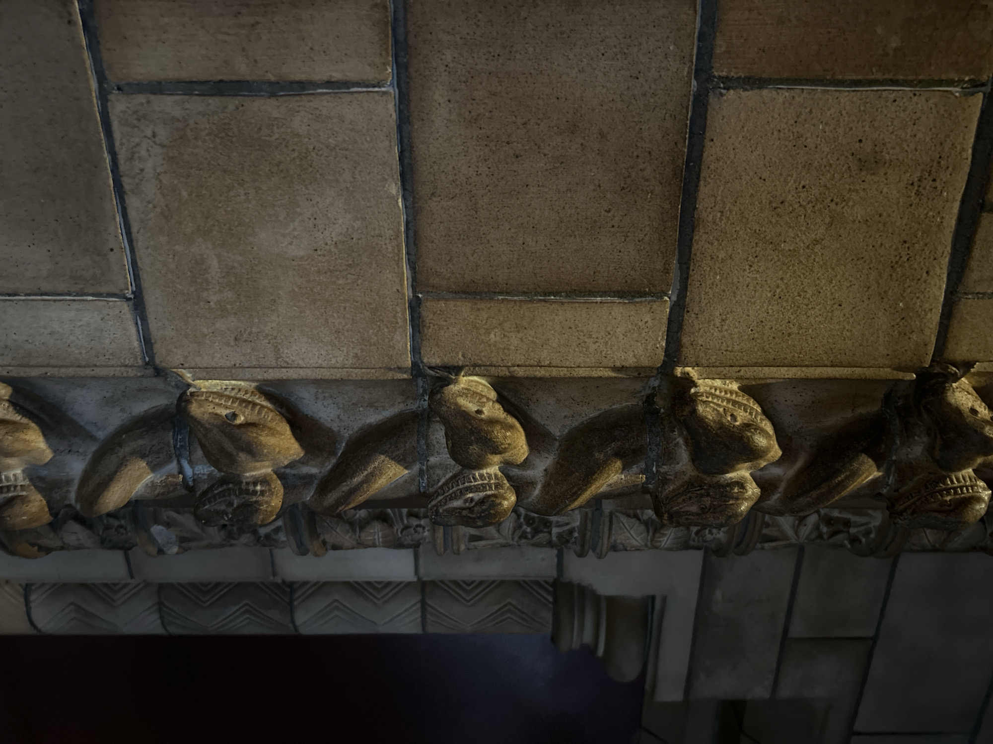 The image shows a close-up of architectural carvings on a stone pillar, likely part of a historic building in London, United Kingdom. The pillar is adorned with a series of grotesque or monstrous faces carved in relief, interspersed with floral or vegetal motifs. The stone appears weathered and aged, with warm, artificial lighting highlighting the intricate details of the carvings. To the left of the pillar, a dark, indistinct background suggests the interior of a building, possibly a museum or a cathedral, with other architectural elements visible in shadow, including what appears to be a column with a decorative base and a chevron pattern on the wall. The main focus is on the detailed texture and form of the carved stone, showcasing the craftsmanship of the period.