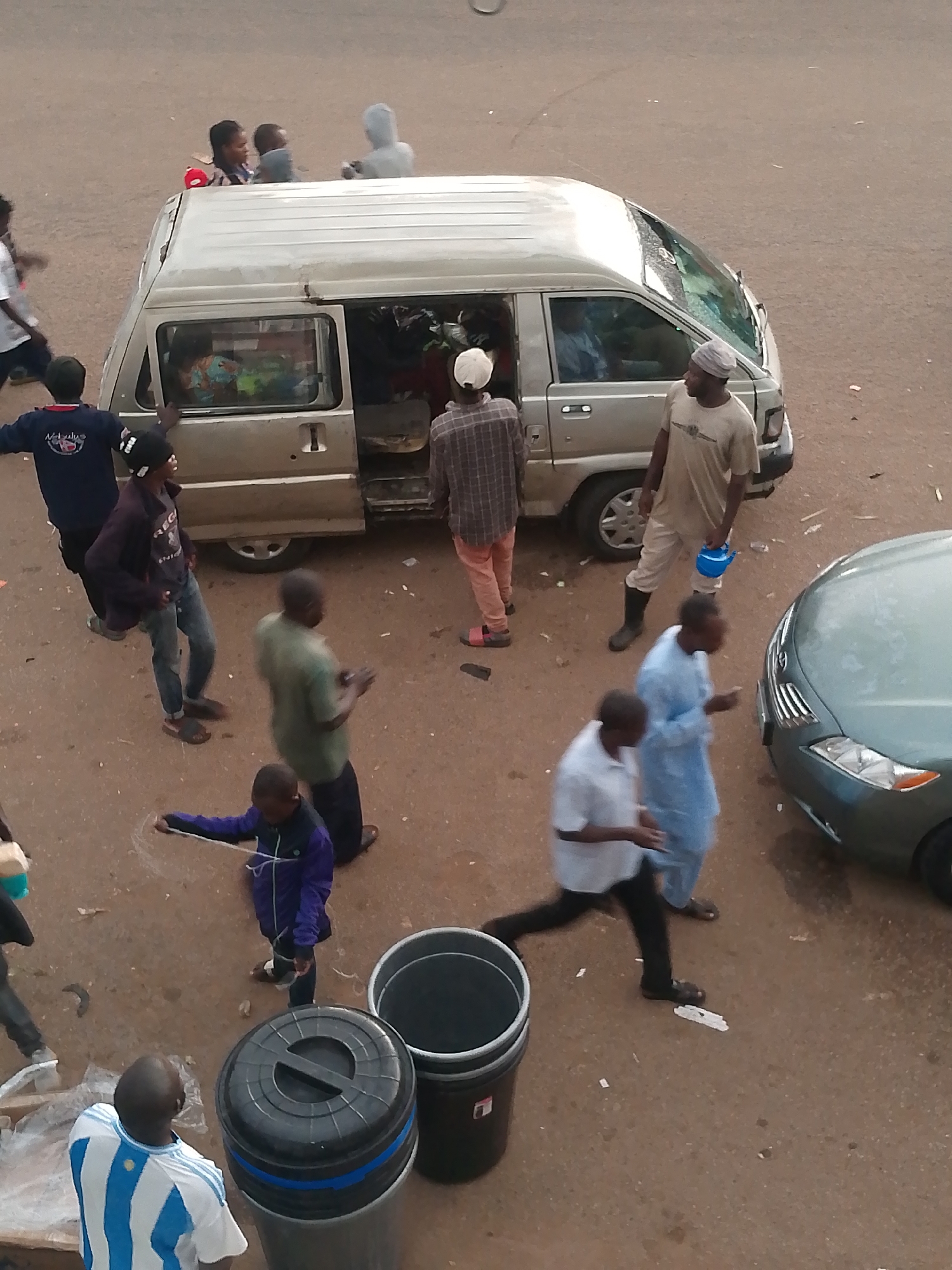 This image depicts an outdoor scene in Bukuru, Nigeria, focusing on a group of individuals gathered around a light-colored van and a light green sedan. The ground appears to be dirt or gravel, typical of an unpaved road or market area.
In the foreground, a person wearing a blue and white striped shirt is seen from behind, facing away from the camera. To their left, a large black trash bin with a ribbed lid is positioned next to a similar gray bin.
Further into the scene, a young person in a purple jacket is holding a white string or rope, appearing to be drawing or playing on the ground. They are wearing dark pants and sandals.
Several adults are present around the van. One person in a light blue garment is walking, their hands outstretched. Next to them, a person in a light-colored shirt and pants, possibly a vendor, is standing and holding a bright blue watering can. A person in a plaid shirt and pink pants is walking away from the van, and another individual in a tan shirt and pants stands near the van's open door.
Inside and around the van, various items are visible, suggesting it might be used for transportation of goods or passengers. People are seen looking into the van or standing near its open side door. One person, wearing a blue shirt and dark pants, is standing with their back to the camera, looking towards the van. Another person in a brown and white checked shirt stands near the open passenger door. A person in a gray shirt and cap is seated on a ledge or step of the van.
In the background, a few individuals are seen standing near the top of the van, one appearing to hold a child. Another person in a hooded gray garment is also visible.
The light green sedan is parked to the right of the frame, with its front end visible. The overall atmosphere appears to be that of a bustling public space, possibly a market or transportation hub.