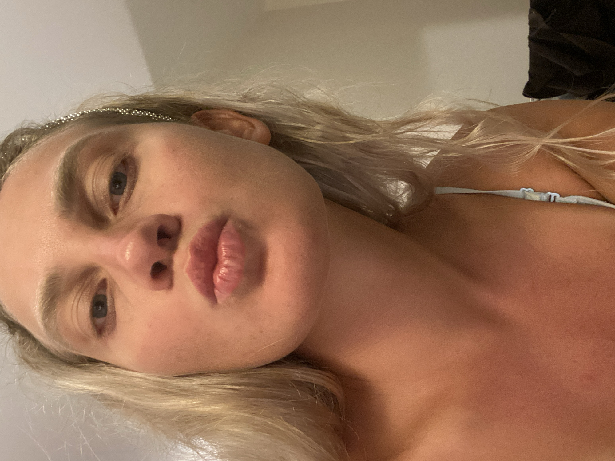 A close-up, low-angle shot shows a woman with blonde hair pursing her lips for a kiss, her head tilted slightly forward. Her eyes are open, and she appears to be looking directly at the camera. A sparkly headband adorns her hair. The background is out of focus, but appears to be an indoor setting with a plain wall. The woman is wearing a light blue top with a thin strap. The lighting in the image is warm and seems to be coming from the front. The overall impression is that of a casual selfie. The city of Aksu, Turkey, is not depicted in this image.