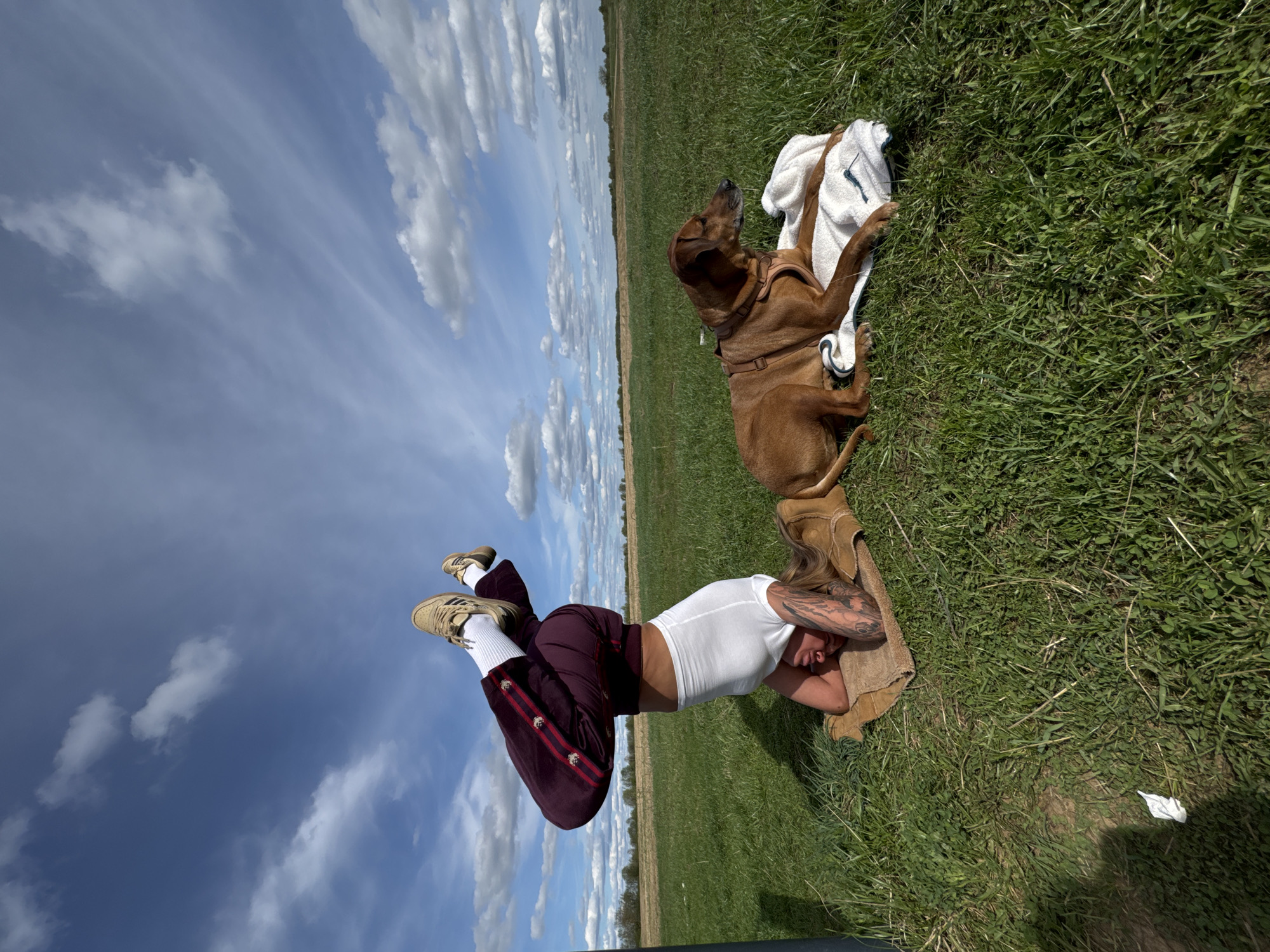 On a grassy field in Bol'shoye Kozino, Russia, a light-skinned person with blonde hair and tattoos lies prone on a brown towel. The person wears a white tank top and maroon pants with red stripes and white buttons. Tan Adidas sneakers with white socks are visible. Adjacent to the person, a brown dog wearing a harness rests on a white towel. The sky is blue with scattered white clouds.