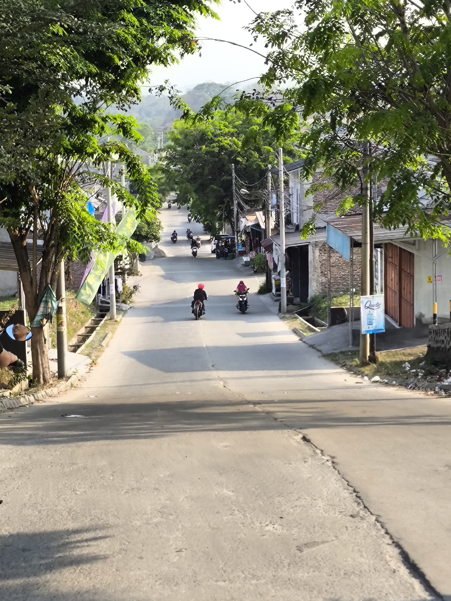A perspective shot looking down a sloped, paved road lined with trees and buildings. On the left, lush green trees arch over the road, with their branches partially obscuring the view. A power pole with wires stands tall on the left side of the road. The road itself has a distinct crack running down its center. Several motorcycles are seen on the road; in the foreground, a motorcycle with a rider wearing a red helmet is heading away from the viewer. Further up the road, another motorcycle with a rider in pink is moving towards the viewer. In the distance, more motorcycles and a tuk-tuk are visible, suggesting a busy street. The buildings lining the road appear to be small shops or houses, some with corrugated iron roofs. On the right side of the road, a white sign with blue text that reads "Quelle" is attached to a pole. The environment appears to be a moderately populated area with a mix of natural elements and urban infrastructure. This scene is located in the city of Cilegon, Indonesia, likely a residential or commercial street given the presence of buildings and local traffic.