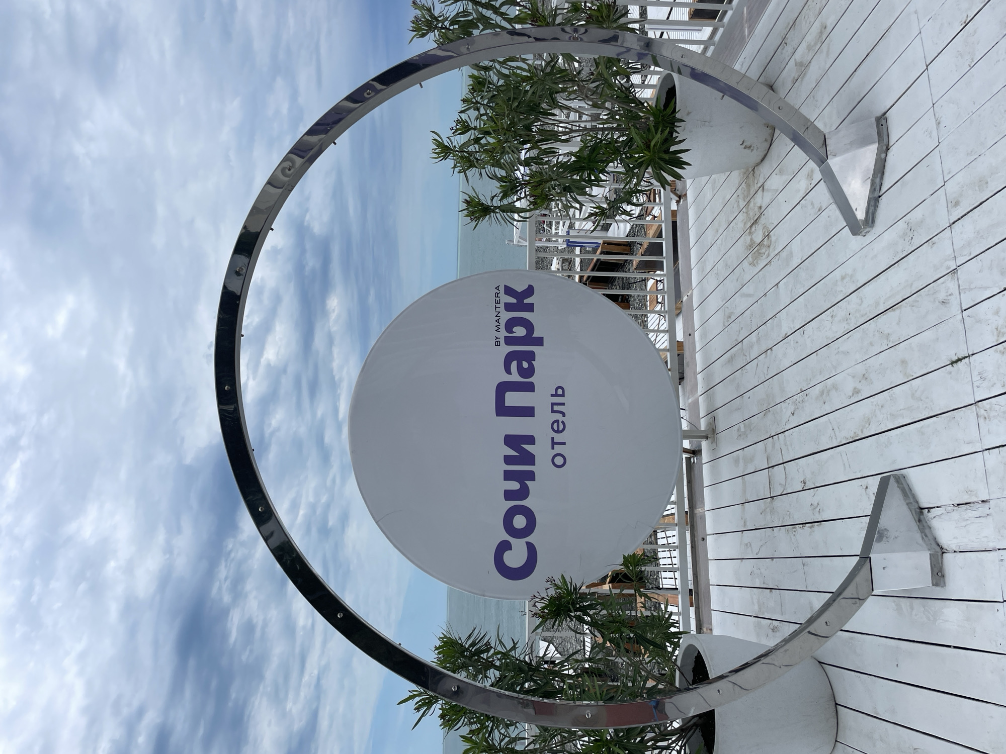 The image shows a circular sign for the "Sochi Park" hotel, located in Sochi, Russia. The sign is primarily white with the hotel name in purple Cyrillic letters. The sign is encircled by a shiny, silver, round metal frame. The sign and frame are mounted on a white wooden deck. Behind the sign, the sea and a cloudy sky are visible. Plants in white pots flank the sign. The scene is oriented in a portrait style.