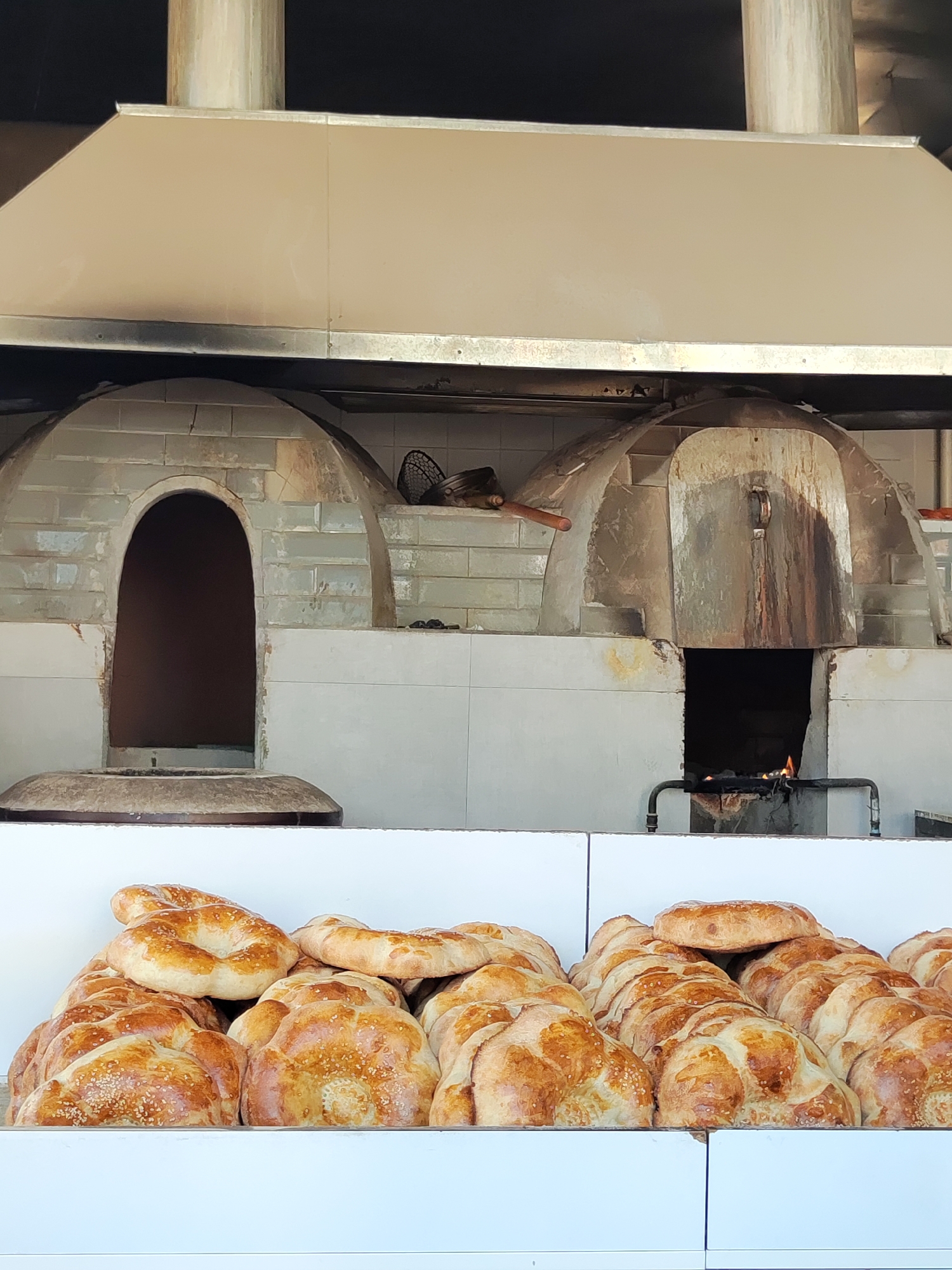 A still shot of a bakery in Tashkent, Uzbekistan, showcases two traditional tandyr ovens. The ovens are dome-shaped, constructed from light-colored bricks and cement, with metal hoods above them. The left oven is open, revealing a dark interior. The right oven has a metal door and a visible opening at the bottom where flames can be seen. In front of the ovens, a display counter is filled with freshly baked bread. The bread loaves are golden brown and appear to have sesame seeds sprinkled on top. The environment suggests a food preparation area, likely an outdoor or semi-outdoor setting given the open-air feel and the presence of industrial-style ventilation.