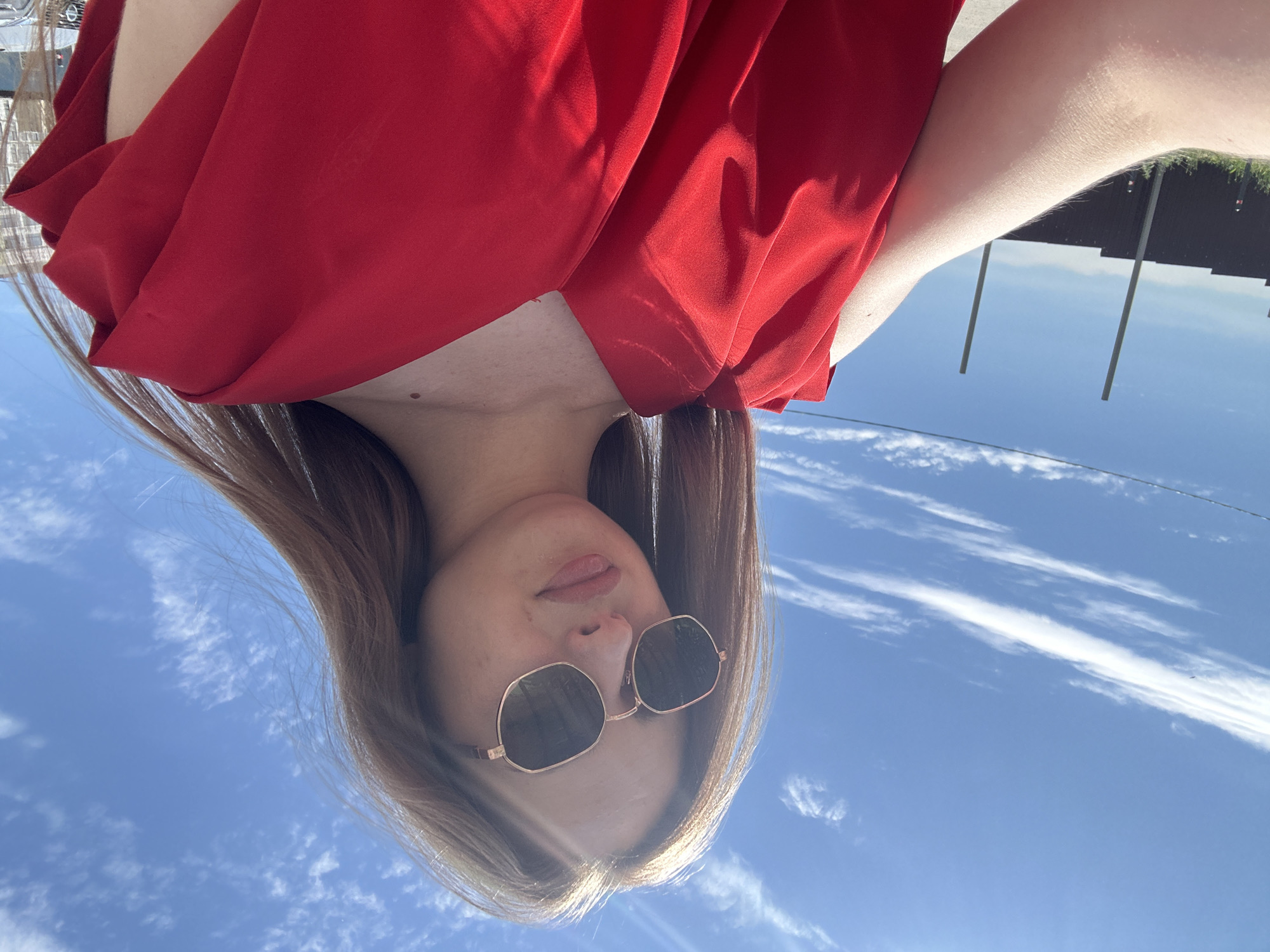 A young woman with long brown hair and sunglasses poses for a selfie outdoors under a bright blue sky with scattered white clouds. She is wearing a vibrant red dress with ruffled sleeves. The background shows a dark fence, two tall poles, and a hint of buildings in the distance. The lighting suggests it is daytime.