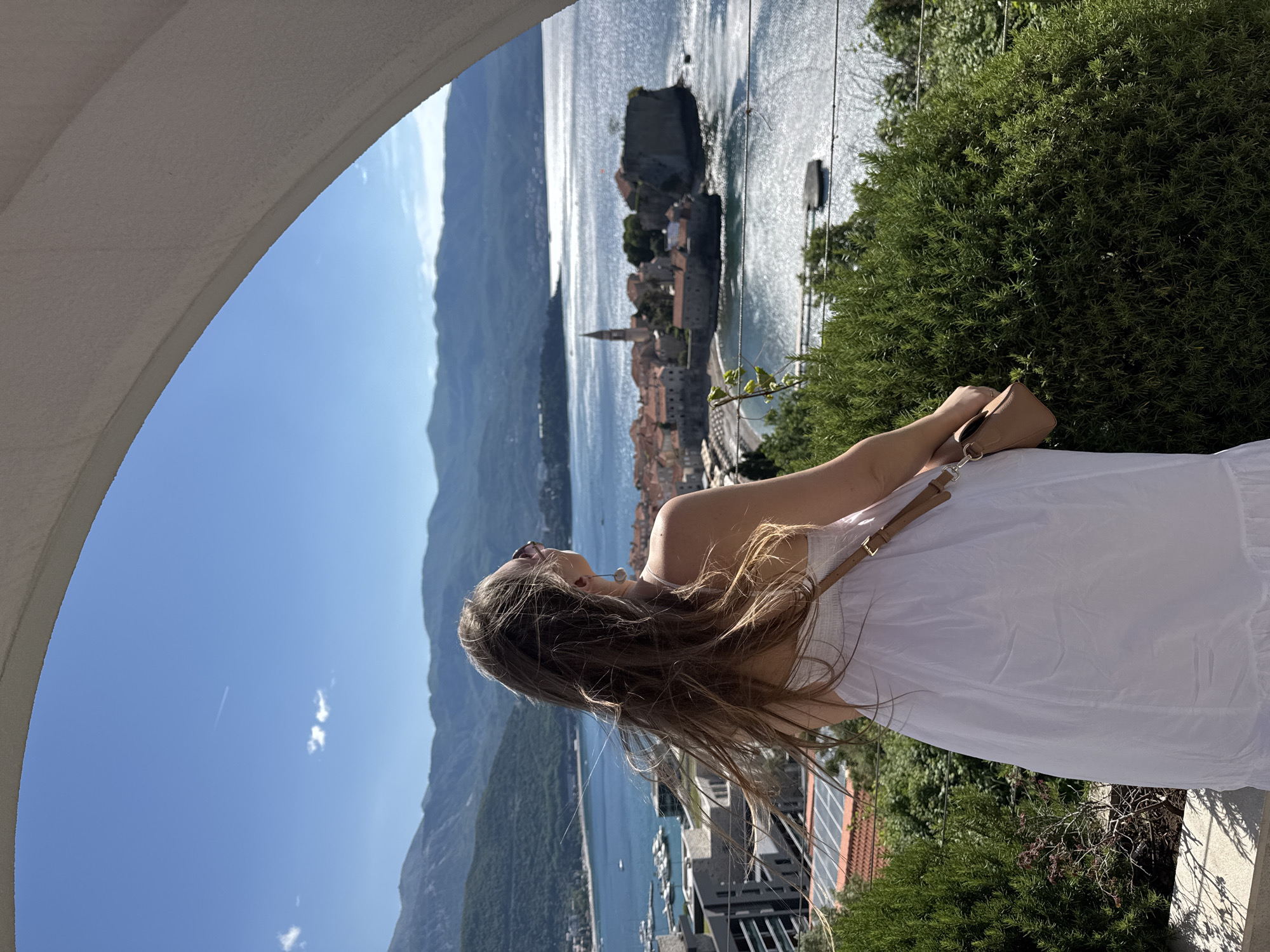 A woman in a white dress gazes out at the Adriatic Sea from a vantage point in Budva, Montenegro. She is framed by a white arch, looking towards the old town with its distinct red rooftops and a church steeple. Mountains rise in the background, and the sea is dotted with boats. There is a large green bush to her right, and buildings and the coastline can be seen below.