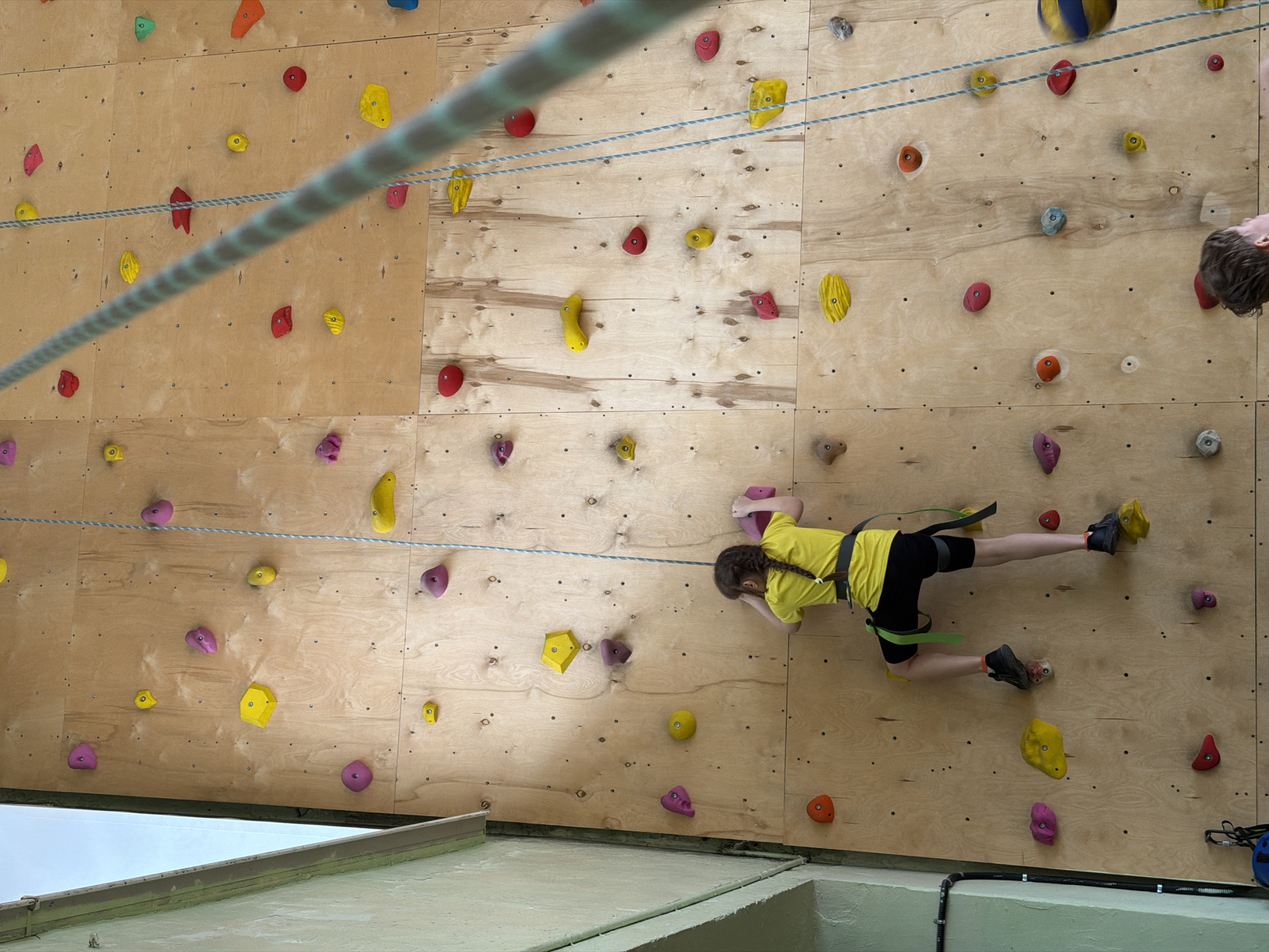 A girl is climbing an indoor rock climbing wall. The wall is made of wood and has various colored holds attached to it. The girl is wearing a yellow shirt, black shorts, climbing shoes, and a climbing harness. She is holding onto the wall with her hands and feet. There is a rope strung across the wall above her. In the upper right corner of the image, a portion of a man’s head can be seen along with a blue and yellow ball. The building where the wall is located is in Izhevsk, Russia.