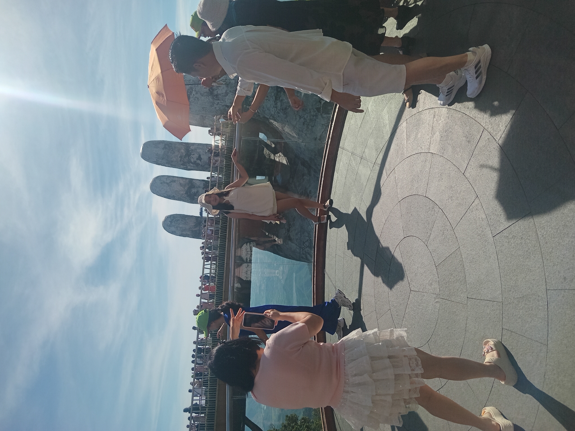 A group of people are gathered on an outdoor viewing platform, likely part of the Golden Bridge in the Bà Nà Hills of Đà Nẵng, Vietnam. The iconic giant stone hands of the bridge are visible in the background. In the foreground, a woman in a light pink top and a tiered white skirt is taking a photo with her phone of another woman in a blue outfit and a green hat. To the right, a man in a white shirt and shorts holds an orange umbrella, and a woman in a black dress stands beside him. Other tourists are visible on the bridge railing in the distance. The ground is paved with large, circular stone tiles, and the sky is a bright blue with scattered white clouds. Sunlight streams down, creating distinct shadows on the ground.