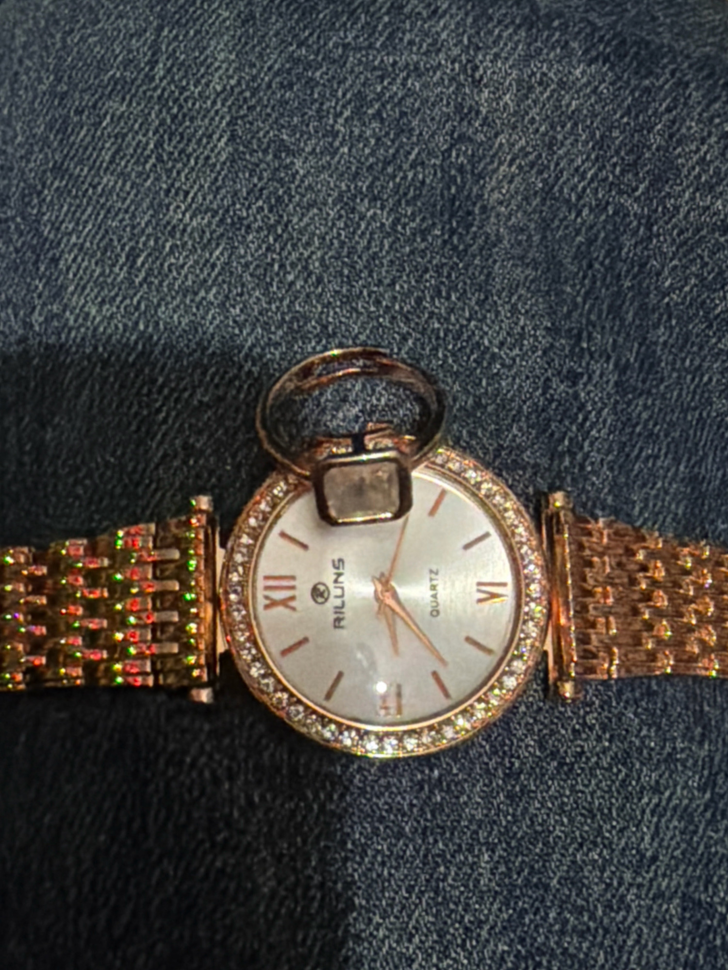 A rose gold analog wristwatch with a diamond-encrusted bezel and a mother-of-pearl face sits on a dark blue denim surface. The watch band is also rose gold and features a woven texture. A ring with a rectangular, clear stone is placed on top of the watch face. The image is a close-up, high-angle shot. There are no people visible in the image. The location in Vapi, India, is not discernible from the visual information provided.