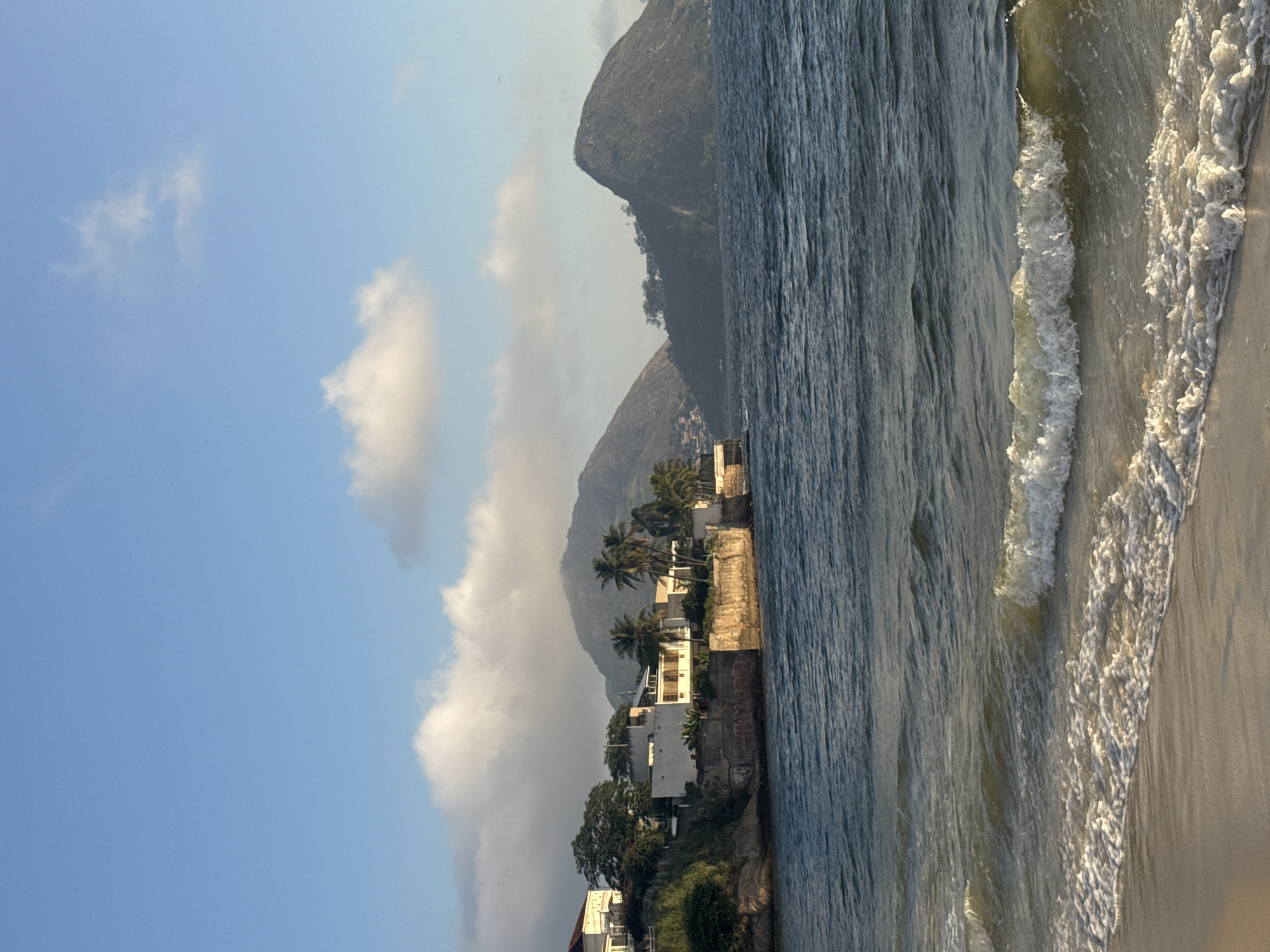 The image captures a coastal view of the city of Niterói, Brazil, specifically the Boa Viagem neighborhood, with the iconic Morro da Urca visible in the background. The foreground features a sandy beach with gentle waves lapping at the shore, creating a foamy white line. The water is a deep blue, reflecting the sky.
Beyond the beach, a rocky coastline is lined with several modern white buildings, some with balconies and terracotta-tiled roofs. Lush green vegetation, including palm trees, grows along the clifftop, contrasting with the man-made structures. The architecture suggests a residential area.
In the mid-ground, a series of imposing, tree-covered mountains rise majestically. The largest mountain, to the right, appears somewhat rounded, while the one to its left is more elongated and has a settlement of houses cascading down its slopes, indicating the urban spread. The sky above is a clear blue, punctuated by a few fluffy white clouds, suggesting pleasant weather conditions, possibly late afternoon due to the soft lighting. There are no people or visible animals in the scene. The overall impression is one of tranquility and natural beauty combined with urban development.