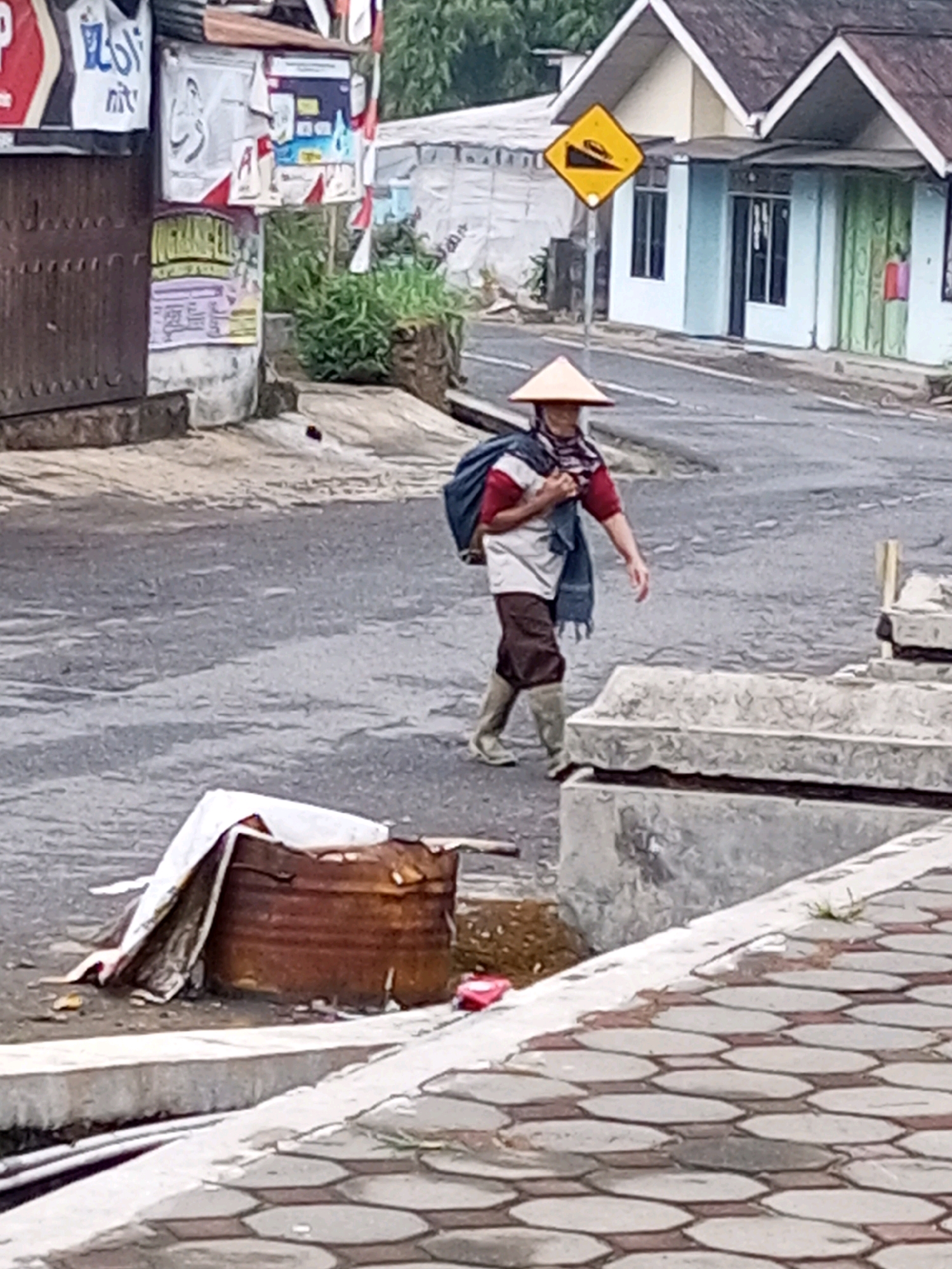 A person wearing a conical straw hat walks down a paved road in Keseneng, Indonesia. The individual is carrying a dark backpack over their shoulders and has a blue scarf draped around their neck. They are dressed in a maroon and white shirt, brown pants, and tall brown boots. To the left of the person, a rusty, cylindrical metal container is covered with a white tarp. Behind the person, a yellow diamond-shaped sign with a black arrow indicating a steep incline is visible on a pole. Several buildings line the opposite side of the road, some with painted facades and open windows or doors. Green foliage is present in the background. The road surface is grey asphalt, and a patterned pavement is visible in the foreground.