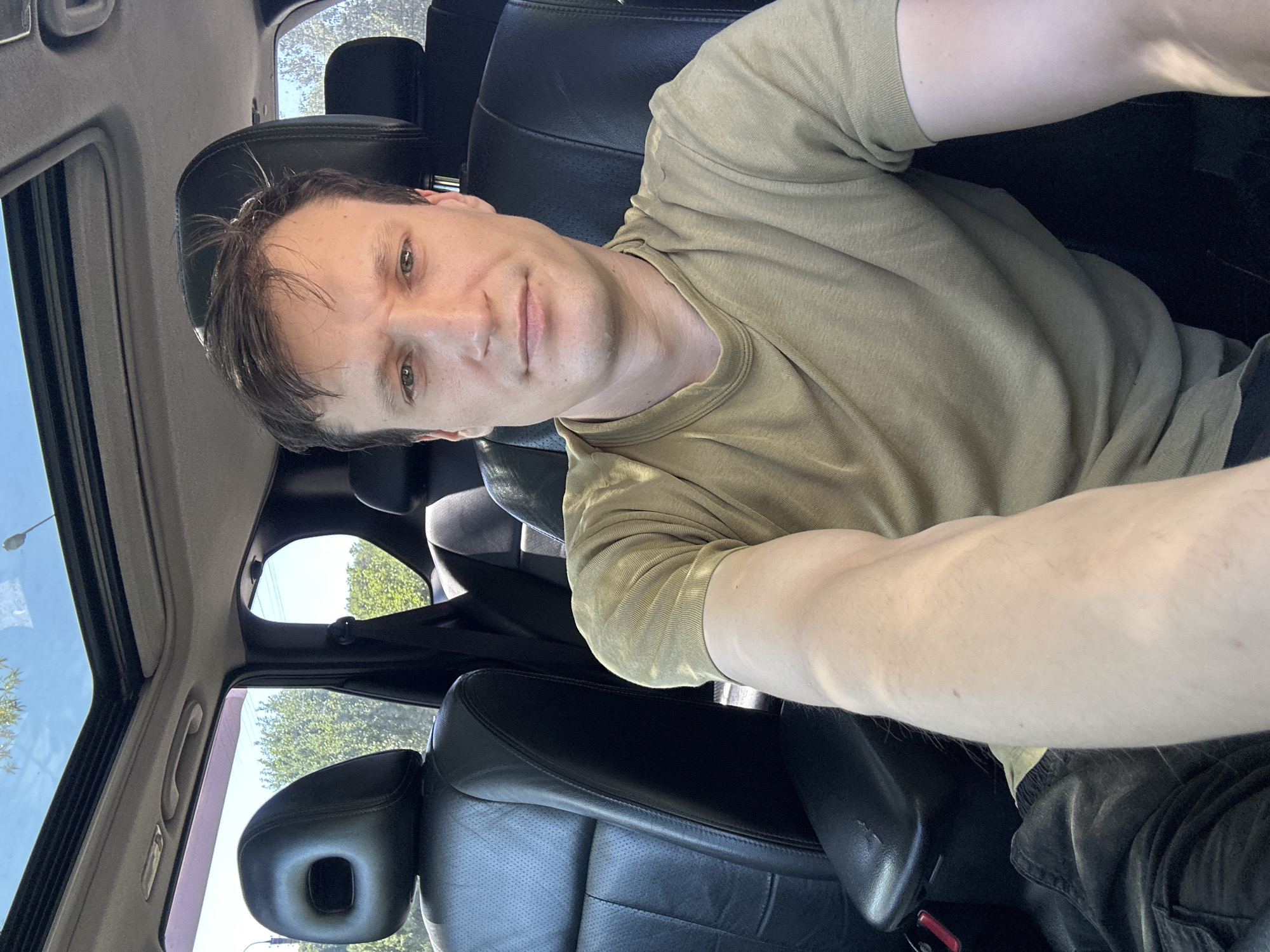A young man with short, dark hair and fair skin is seated in the driver's seat of a car, taking a selfie. He is wearing a plain olive-green t-shirt. The interior of the car is visible, with black leather seats and a sunroof open to a bright, sunny sky. Trees and greenery can be seen through the open sunroof and windows, suggesting an outdoor location, possibly on the outskirts of Makiv, Ukraine, given the context. The man's expression is neutral. The camera angle is from his perspective, looking slightly down at himself.