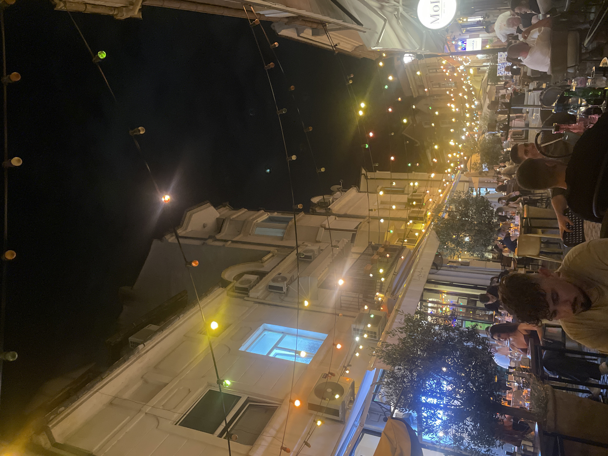 This image captures a nighttime scene on a bustling pedestrian street in the city of Unknown, Unknown. The street is lined with multi-story buildings, predominantly light-colored, featuring windows and multiple exterior-mounted air conditioning units. Overhead, numerous strands of colorful string lights, displaying yellow, orange, green, and red bulbs, are strung across the thoroughfare, providing primary illumination.
The street is densely populated with dozens of individuals seated at outdoor tables, indicative of a lively dining or social establishment. In the foreground, a man with curly dark hair and a beard is seated, facing away from the camera. To his left, another man with facial hair looks towards the camera. A woman with dark hair, wearing a reddish top, is seated at a table on the left mid-ground. Many tables feature glassware, bottles (including wine bottles), and visible shisha (hookah) pipes. People are engaged in conversation, eating, and drinking. Waitstaff are also visible, including a man in a white shirt standing near a circular sign on the far right that displays "Mol" and "EST 2021".
Potted trees and shrubs are interspersed among the tables, providing natural elements. Awnings extend over some seating areas, one on the left displaying partial text resembling "Luba". In the background, additional establishments with outdoor seating and more string lights extend down the street. One building in the mid-ground features a blue-and-white sign with text that appears to read "Junias". The sky above is dark, indicating night.