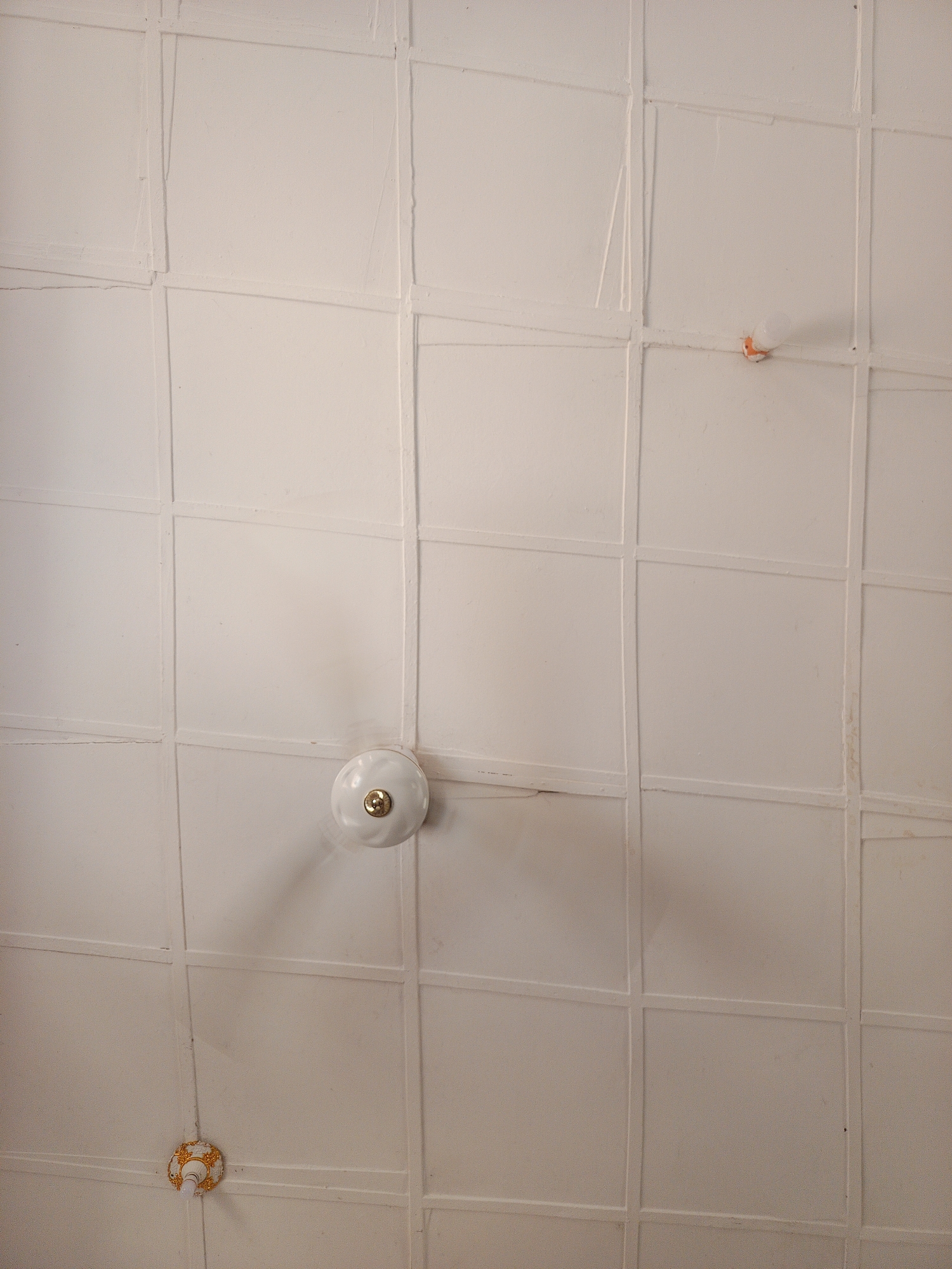 The image displays a detailed, upward-facing view of a white paneled ceiling located in Anguwar Jaba, Nigeria. The ceiling is constructed from numerous off-white, slightly textured square or rectangular panels, arranged in a grid pattern. The seams between the panels are demarcated by raised, painted lines, exhibiting minor irregularities and evidence of wear, including subtle cracks and minor discoloration.
A three-bladed white ceiling fan, with a central gold-colored cap, is centrally positioned in the lower middle section of the frame and appears to be in motion, indicated by the blurred appearance of its blades.
In the upper right quadrant of the ceiling, a bare, frosted white light bulb is affixed directly to a panel via a visible orange-colored socket. In the lower left quadrant, a second light fixture is present. This fixture features a smaller, clear light bulb emerging from an ornate, circular base decorated with gold-colored scrollwork patterns.
No individuals, discernible actions, or interactions are present within the frame, which solely focuses on the architectural elements and fixtures of the ceiling.