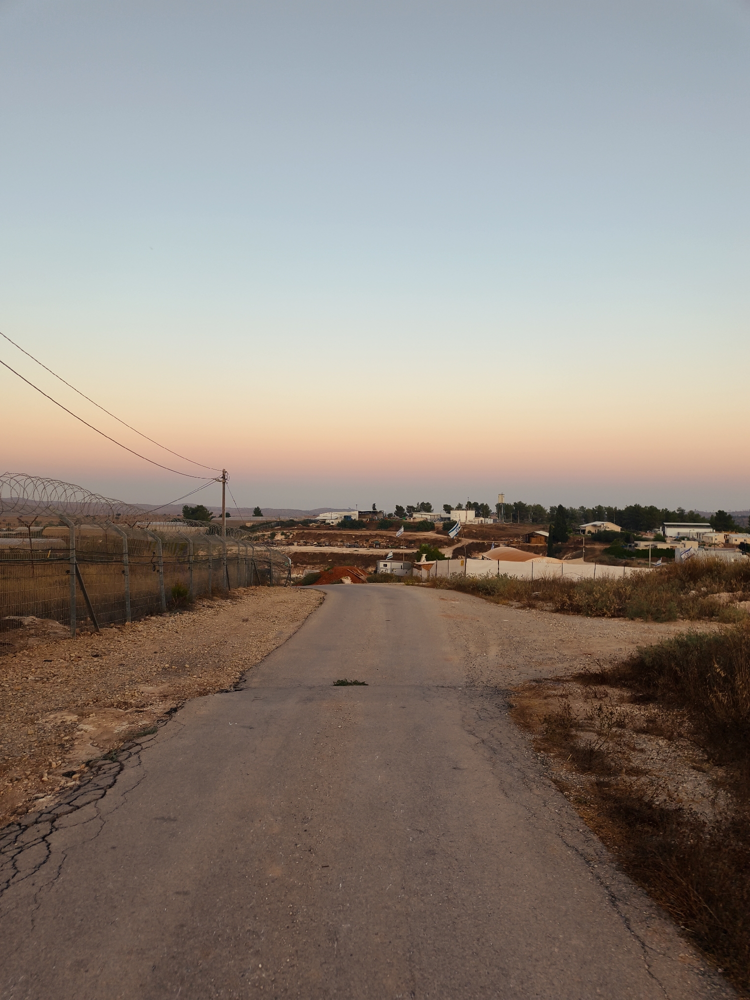 The image shows an asphalt road leading into a settlement in the Lachish Regional Council, Israel, during sunset. The sky displays a gradient of light blue and soft pink hues. To the left of the road, a chain-link fence topped with barbed wire runs along the perimeter, separating the road from a dry, rocky terrain with sparse vegetation. Utility poles and wires are visible near the fence. In the background, the settlement consists of low-rise buildings, some with white roofs, scattered among trees and dry brush. Several Israeli flags are planted within the settlement. The road itself is cracked and weathered, with patches of gravel and dried grass on its surface and edges. The overall scene conveys a sense of an arid, rural environment at dusk.