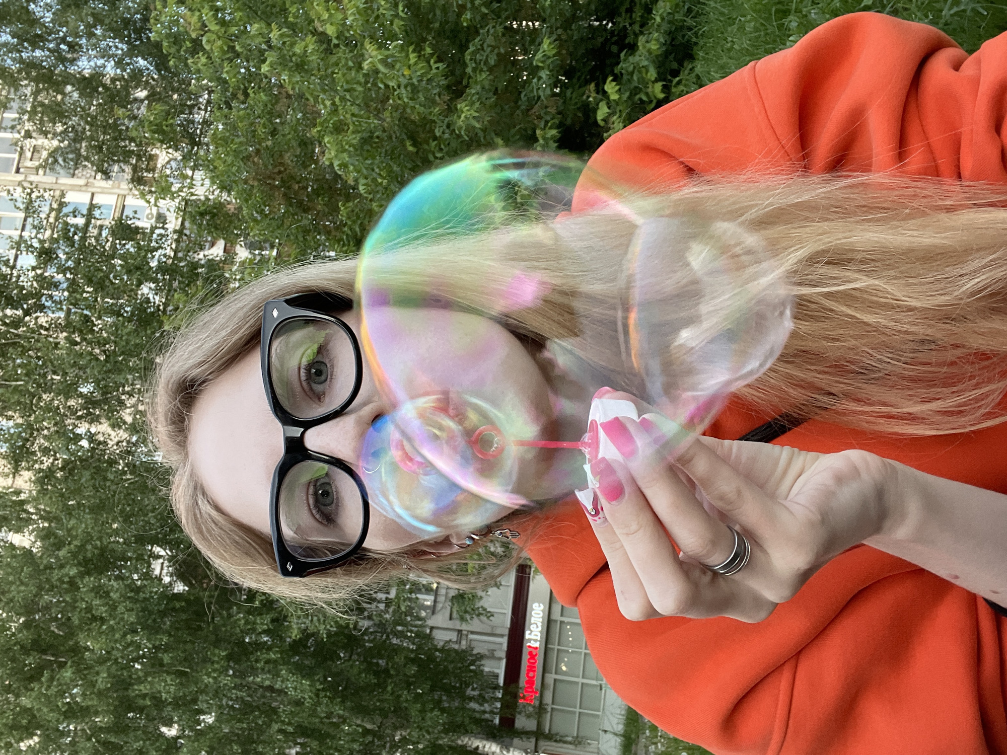 A close-up, rotated image shows a person, likely female, with blonde hair and black-framed glasses, in the process of blowing iridescent soap bubbles. Their left eye is visible through the glasses, reflecting green foliage. They wear a bright orange hoodie. Their right hand, adorned with long, pink and white decorated nails, holds the bubble wand near their mouth, from which several colorful, shimmering bubbles emanate. A silver ring is visible on their left ring finger.
The background consists of lush green trees and a multi-story building, suggesting an urban outdoor setting. On the building, a prominent red sign with white lettering reads "Красное&Белое" (Red&White), a well-known Russian retail chain, and another smaller sign with "home" is partly visible. The lighting indicates it is daytime, possibly late afternoon, with clear weather. The overall scene conveys a playful and whimsical moment in what appears to be a park or street in Izhevsk, Russia.