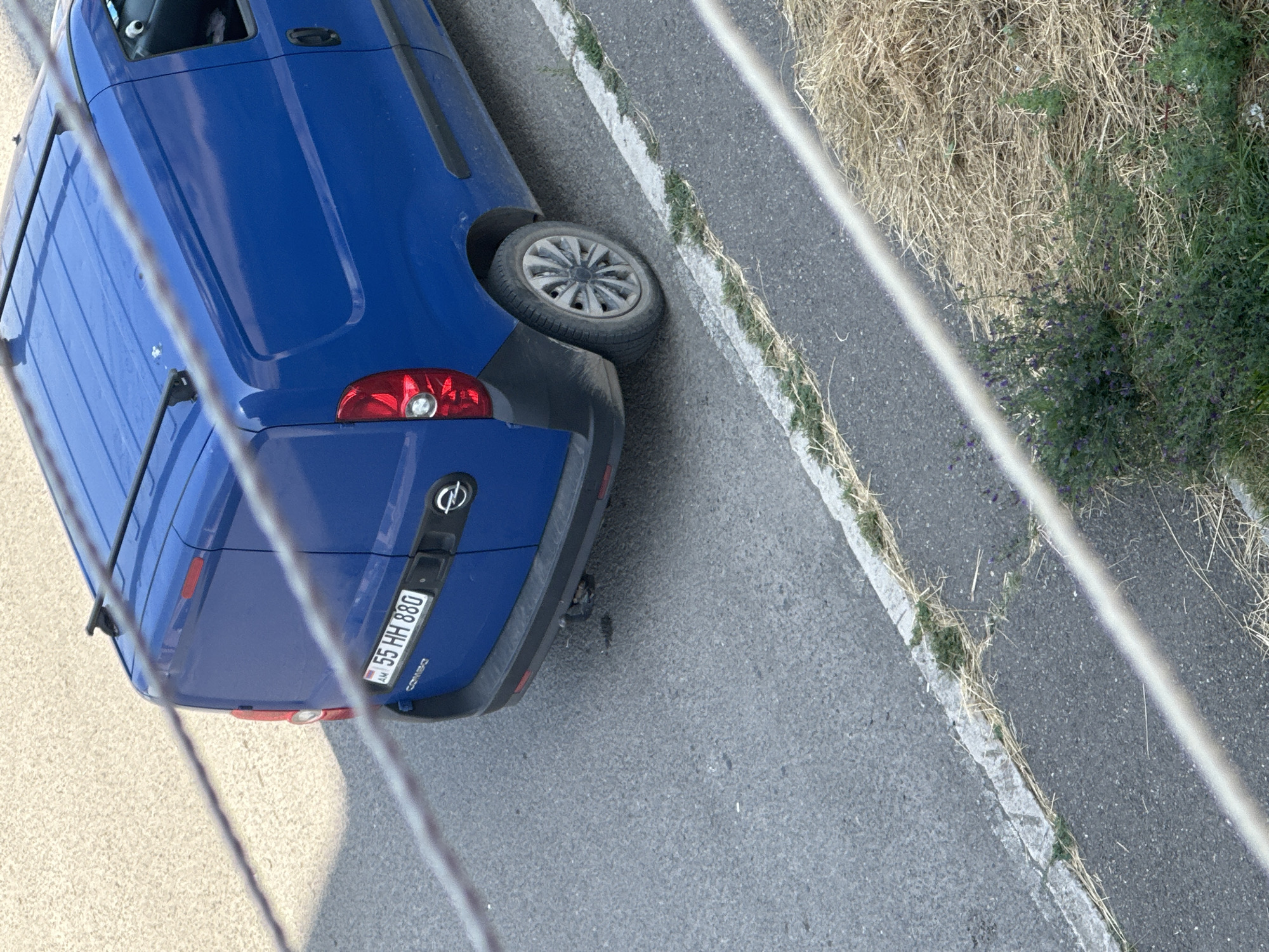 The image shows a blue Opel Combo van parked on an asphalt road in Gyumri, Armenia. The van has a license plate that reads "AM 55 HH 880". The van is facing away from the camera and is partially obscured by two diagonal wires that run across the foreground. The road has a white line marking its edge, and beyond that is a strip of dry grass and some green vegetation with small purple flowers. The background is a textured gray surface, likely more asphalt or pavement. The lighting suggests it is daytime.