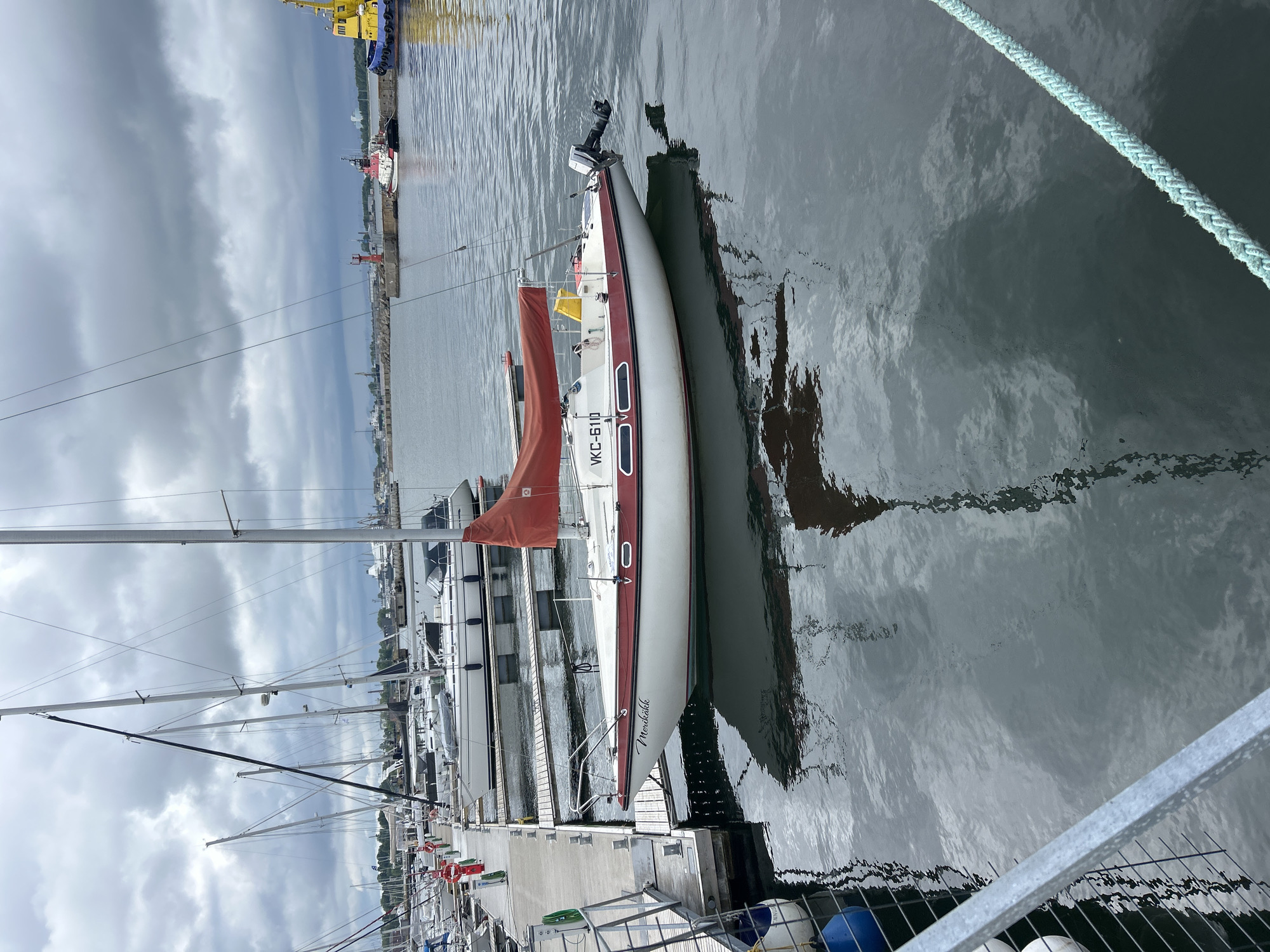 The image shows a sailboat docked at a marina. The sailboat, named "Merikakk" and marked with "VKC-6110", has a white hull with a red stripe and a red sail. It is moored to a wooden dock. In the background, other sailboats and larger vessels are visible. The sky is overcast with dark clouds. The location is identified as Tallinn, Estonia, likely in a harbor area. There are no people visible in the immediate foreground or on the sailboat. The water is calm, reflecting the sky and the boats. The overall scene depicts a quiet day at the marina.