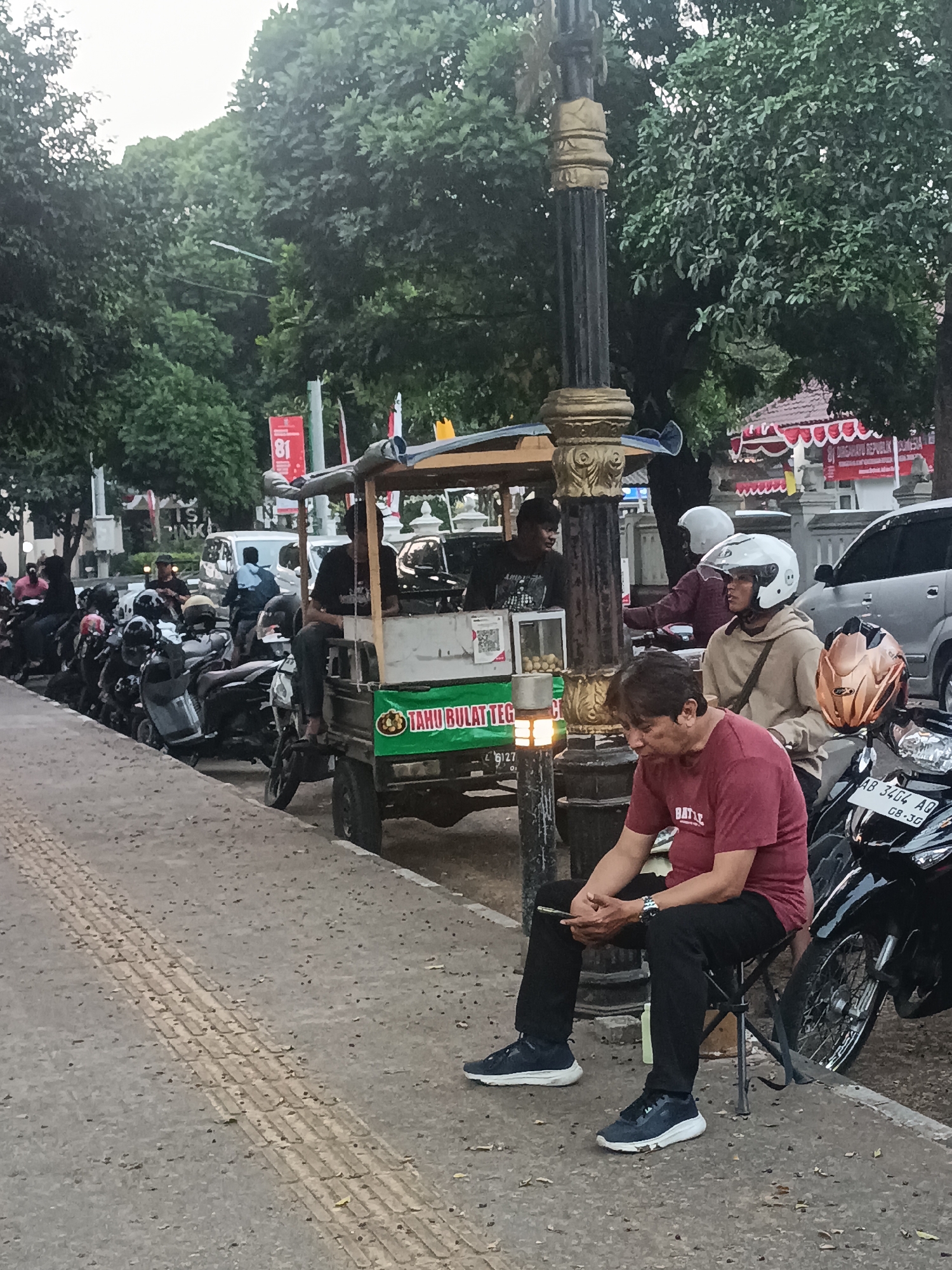 A medium shot captures an outdoor scene in Sleman, Indonesia, featuring a man sitting on a folding stool and looking at his phone. He is wearing a maroon t-shirt with white lettering, black pants, and dark sneakers. To his right, a black motorcycle is parked, and its license plate is partially visible, reading "AB 3404 AQ". In the background, several other motorcycles are parked along the sidewalk. A food cart selling "Tahu Bulat" is also visible, with a vendor tending to it. Trees and buildings line the street, and a decorative lamppost stands prominently near the food cart. The overall lighting suggests it is daytime, possibly late afternoon or early evening.