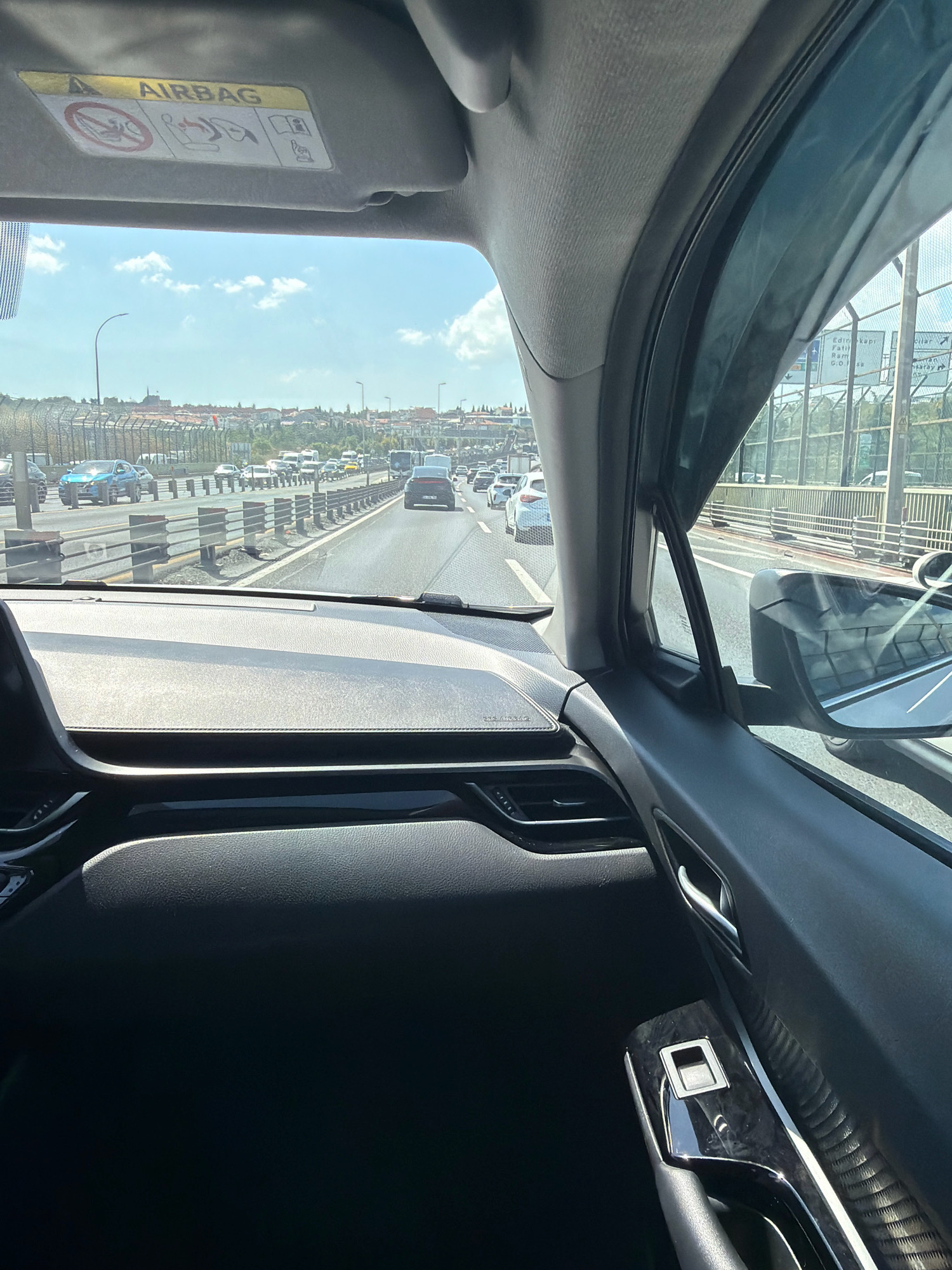 The image shows a view from inside a car, looking out at a traffic jam on a highway. The car's interior is visible in the foreground, including the dashboard and part of the door. The "AIRBAG" warning label is visible on the sun visor. Outside, there are many cars on the highway, with buildings and trees in the background. The sky is blue with some clouds. The location is identified as Eyüpsultan, Turkey.