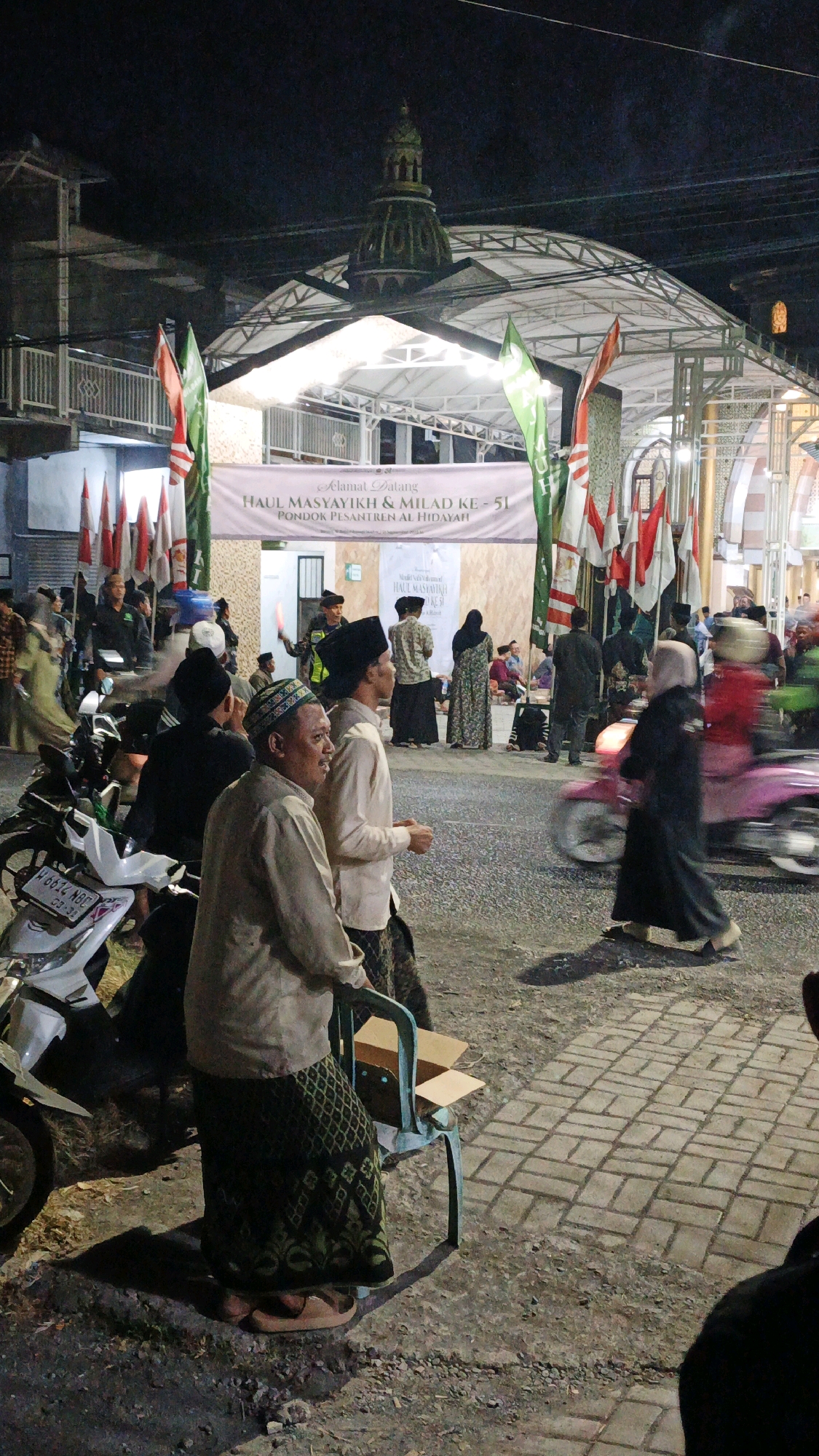The image depicts an outdoor nighttime scene in front of a building adorned with a domed roof and a canopy, possibly a place of worship or a community hall. A banner hangs across the front, reading "SELAMAT DATANG HAUL MASYAYIKH & MILAD KE - 51 PONDOK PESANTREN AL HIDAYAH," indicating an event for the Al Hidayah Islamic Boarding School in Randegan, Indonesia. Numerous flags, some red and white (national Indonesian flags) and others green, are displayed.
In the foreground, several individuals are visible. Two men in the center are looking to the right; one wears a light-colored shirt and a patterned sarong, while the other wears a white shirt and a black cap. A motorcycle is parked to the left of these men. Further back, a group of people are walking and standing. A person on a pink motorcycle is in motion, creating a blur. The ground is paved with bricks. Other elements include a metal chair with an open cardboard box on it. The overall atmosphere suggests a community gathering or religious event.