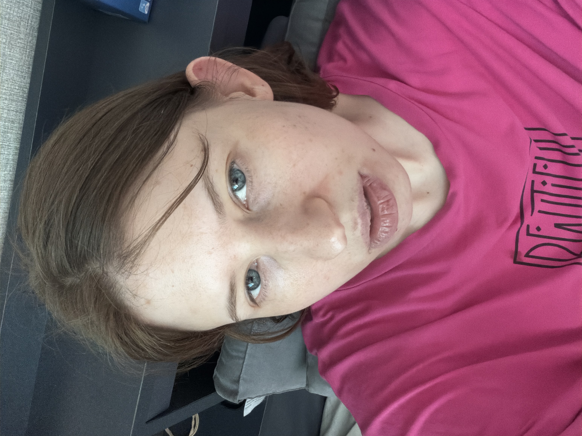 A close-up, high-angle selfie shows a person with fair skin and light brown hair looking into the camera. They are wearing a bright pink t-shirt with a black graphic design on it. The person has blue eyes, light freckles scattered across their face, and their lips are slightly parted. The background is blurred but appears to be an indoor setting, possibly a bedroom, with a gray headboard or pillow and a dark shelf. The lighting is natural, suggesting daytime. The image does not contain any information about the city of Dongducheon-si, South Korea.