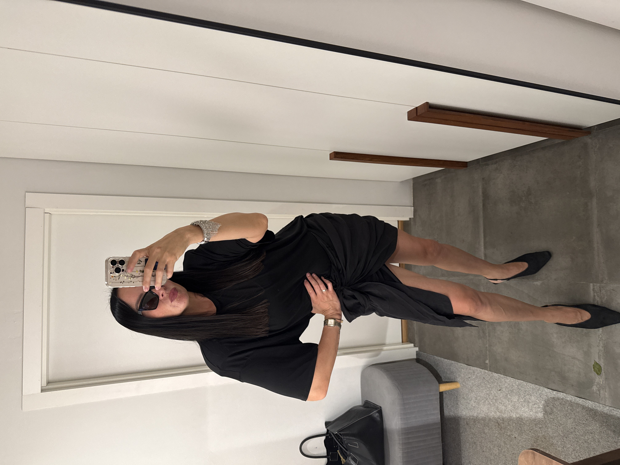 A woman is taking a mirror selfie in what appears to be a fitting room or dressing area. She is wearing a black t-shirt dress with a knotted detail at the waist, black pointed-toe slingback heels, large black sunglasses, and a sparkly bracelet on her left wrist. She is holding a smartphone in her right hand to take the picture. To her left, there is a grey ottoman with wooden legs and a black tote bag on it. The background consists of a white door, a white wall with vertical paneling, and a large mirror. The floor is tiled with large grey tiles. The image does not provide any information that would place it in the city of Chaikovsky, Russia.