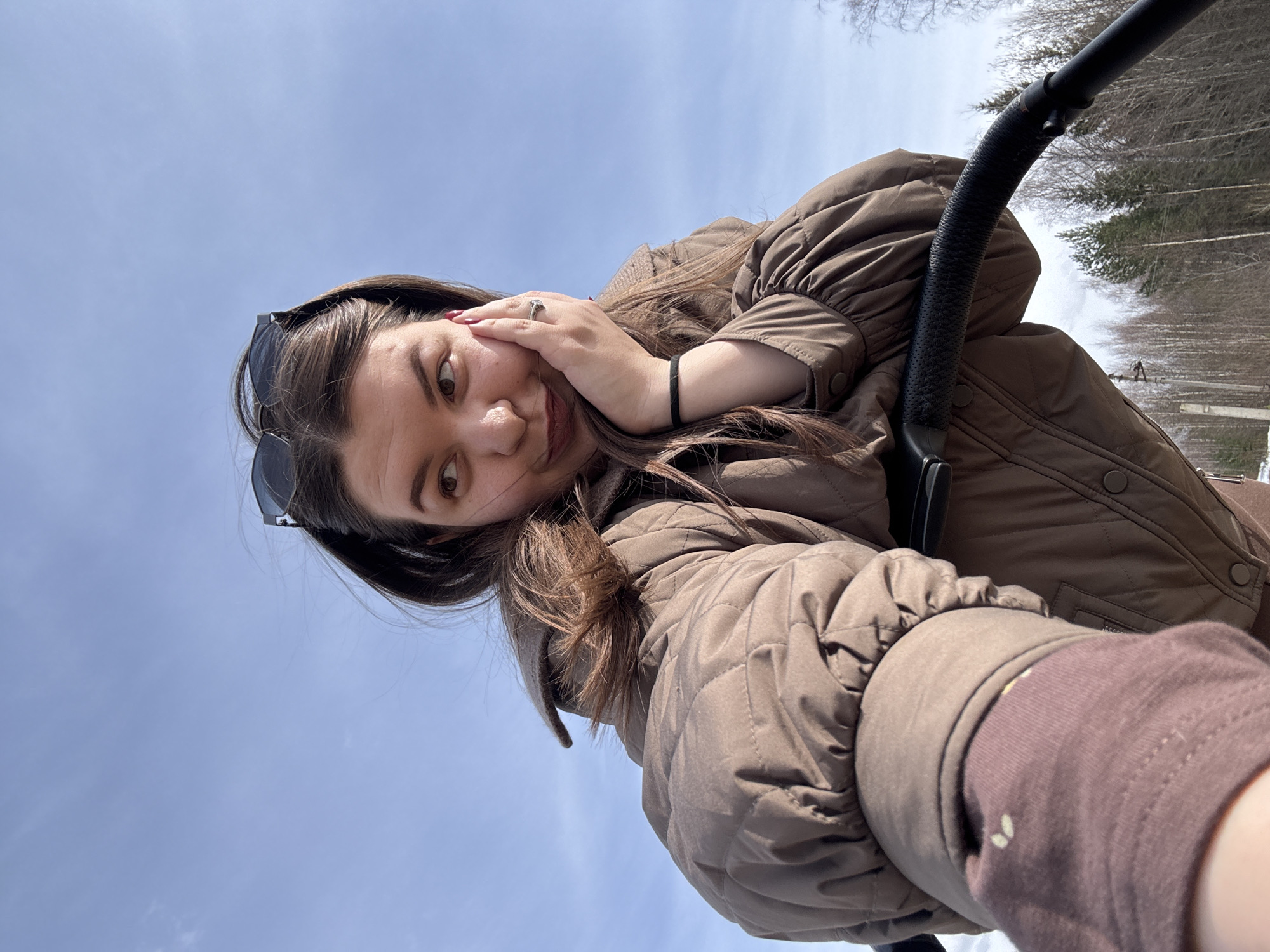 A selfie shows a woman with dark hair and sunglasses perched on her head, wearing a brown puffer jacket, posing on a ski lift. She is holding her hand up to her face in a playful pose. Behind her is a clear blue sky and snow-covered trees in Марковский, Russia. A portion of the ski lift is visible on the right.