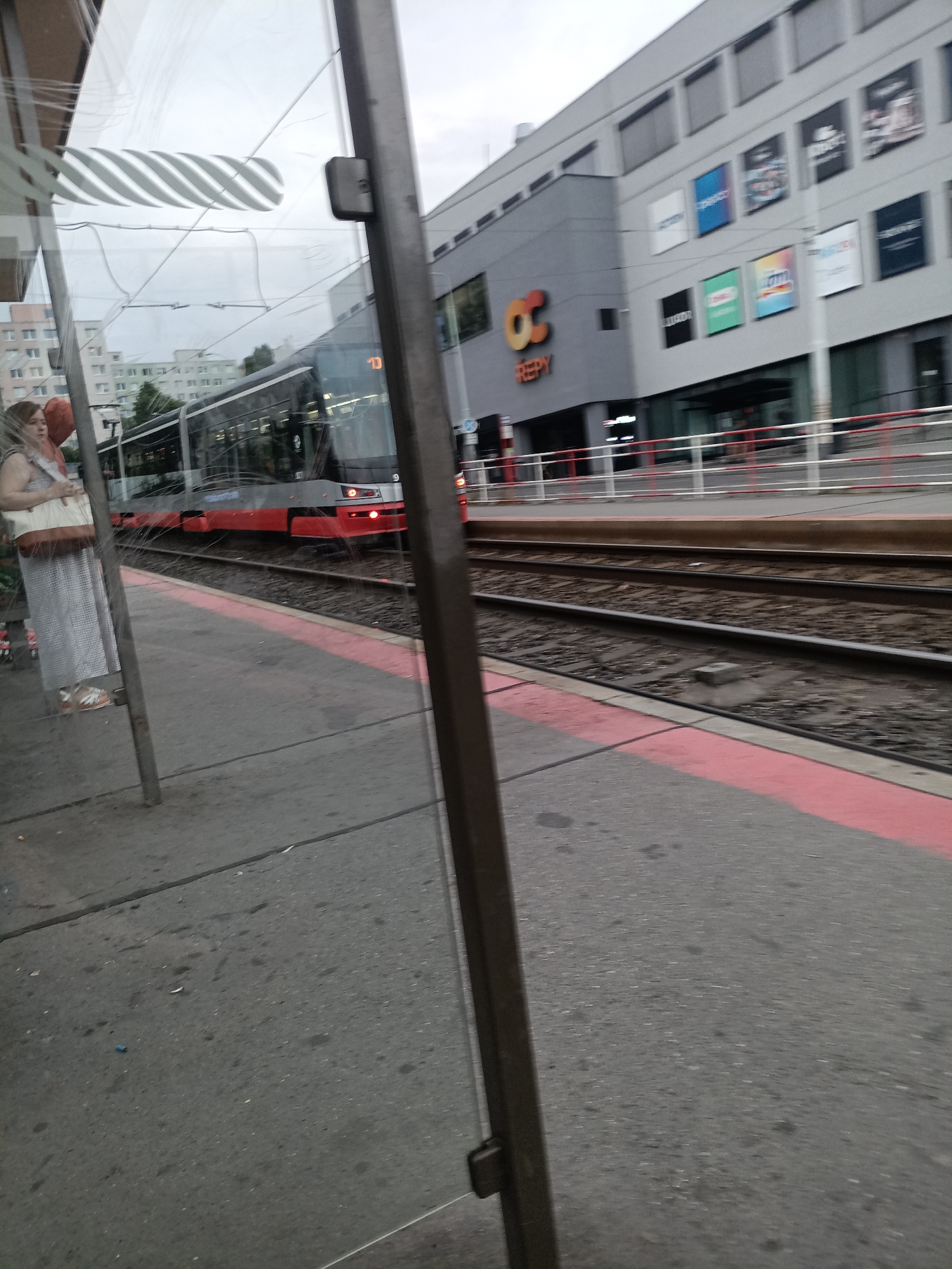 The image shows a tram stop with a tram on the tracks. In the background, a modern building complex with the logo "OC REPY" is visible. A woman stands at the tram stop, holding a bag and an orange object. The tram is red and grey, and appears to be moving or stopped at the station. The tram stop has a grey concrete platform with a red line marking the edge. The overall environment is urban, with buildings, tram tracks, and overhead wires visible. The weather appears to be overcast.