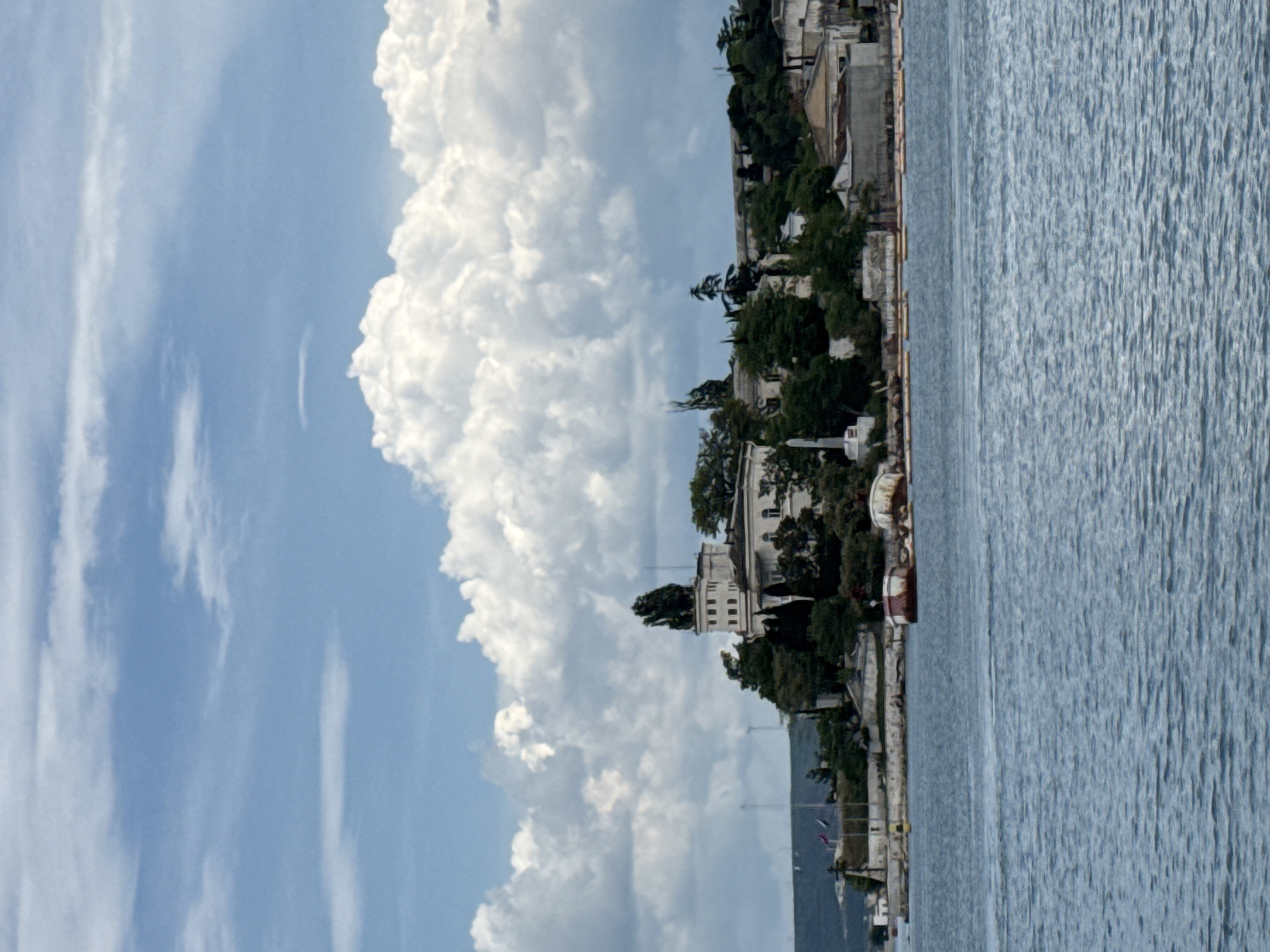 The image displays a panoramic view of a large body of water with a landmass, likely a peninsula or an island, extending into it. The upper portion of the frame is filled with a bright blue sky partially covered by numerous large, white, textured cumulus clouds, with some wispy cirrus clouds visible higher up.
The lower half of the image shows a calm body of water, colored in shades of blue and grey, with minor ripples on its surface.
The central feature of the image is the landmass, which hosts several structures and dense vegetation. On an elevated section of this landmass, slightly left of center, stands a prominent, light-colored building. This building features multi-story construction with architectural details, including what appears to be a tower on its left side, heavily adorned or topped with dense, dark green trees and foliage. To the right of this main building, additional less distinct structures are visible, nestled among trees.
To the left of the main building, closer to the water's edge, a slender, white monument or obelisk on a square base is clearly visible. Further to the left, along the shoreline, several flags on poles are present, though their specific designs are indistinguishable. The shoreline itself appears to be a constructed embankment or retaining wall.
Along the water's edge in the middle ground, several small, flat, yellow or orange objects, possibly small boats or pontoons, are lined up. Further to the right, closer to the shore, two larger, roughly circular or irregular structures, possibly derelict boats or barges with rust-colored elements, are partially submerged or beached.
The landmass is heavily forested with various dark green trees and shrubs, particularly dense around the main buildings, providing a strong contrast to the lighter architectural elements. No people are visible in the scene. The overall scene is brightly lit under natural daylight. This environment is situated within a bay in the city of Unknown, Unknown.