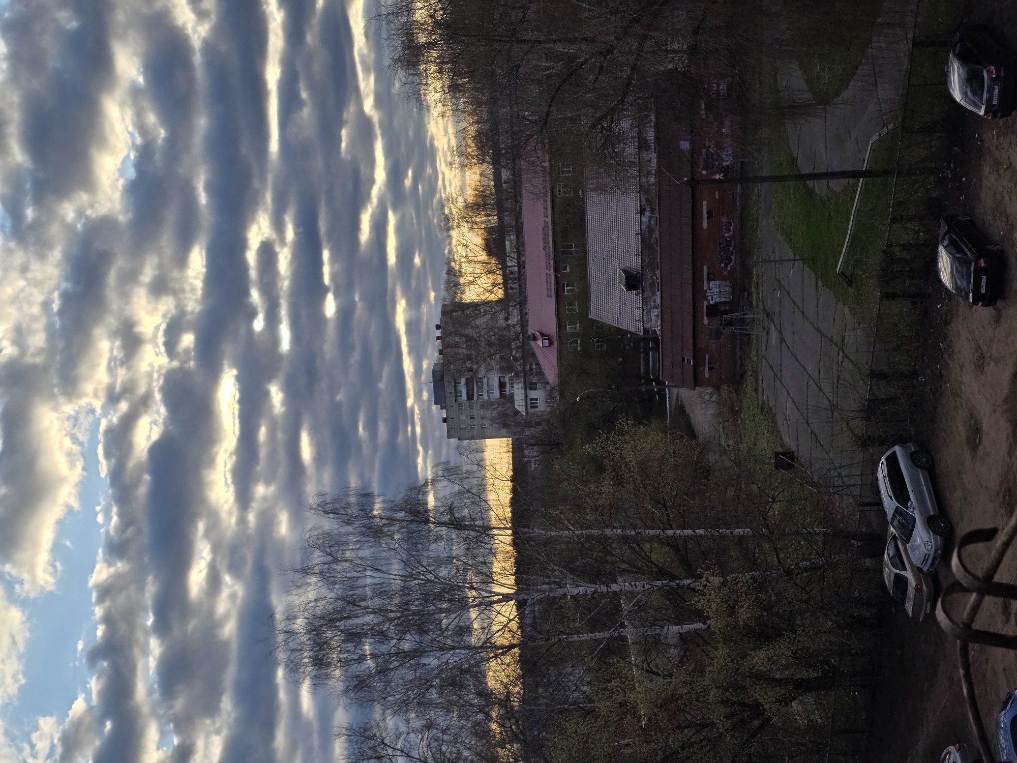 Here's a description of the image:
The image shows an elevated perspective of a neighborhood scene in Chaikovsky, Russia. The sky is filled with altocumulus clouds illuminated by the setting sun, giving them a striated appearance. Below the sky, a mix of leafless and budding trees occupy a middle ground.
In the lower right quadrant, parked cars are visible. To the left, there are a series of buildings with differing roof colors and architectural styles. The ground appears to be a mix of paved surfaces, grassy areas, and bare earth. A fence is also visible.
No people are visible in the image. The overall scene is one of a typical residential area in the evening light.