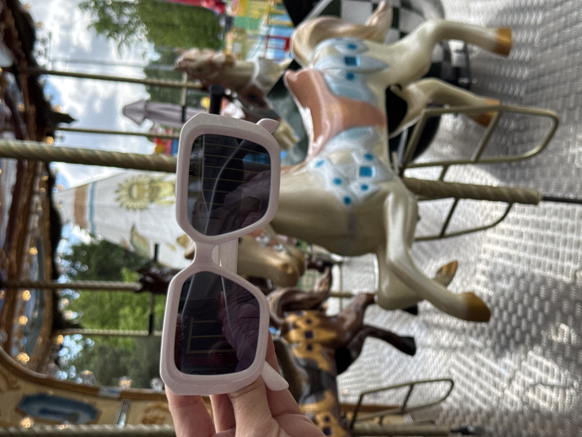A hand holds a pair of white-framed sunglasses in front of a carousel. The carousel features painted horses and other animals. The environment appears to be outdoors, potentially in a park or amusement area. There is a reflection in the sunglasses. Given the presence of a carousel, it is potentially located in Saint Petersburg, Russia.