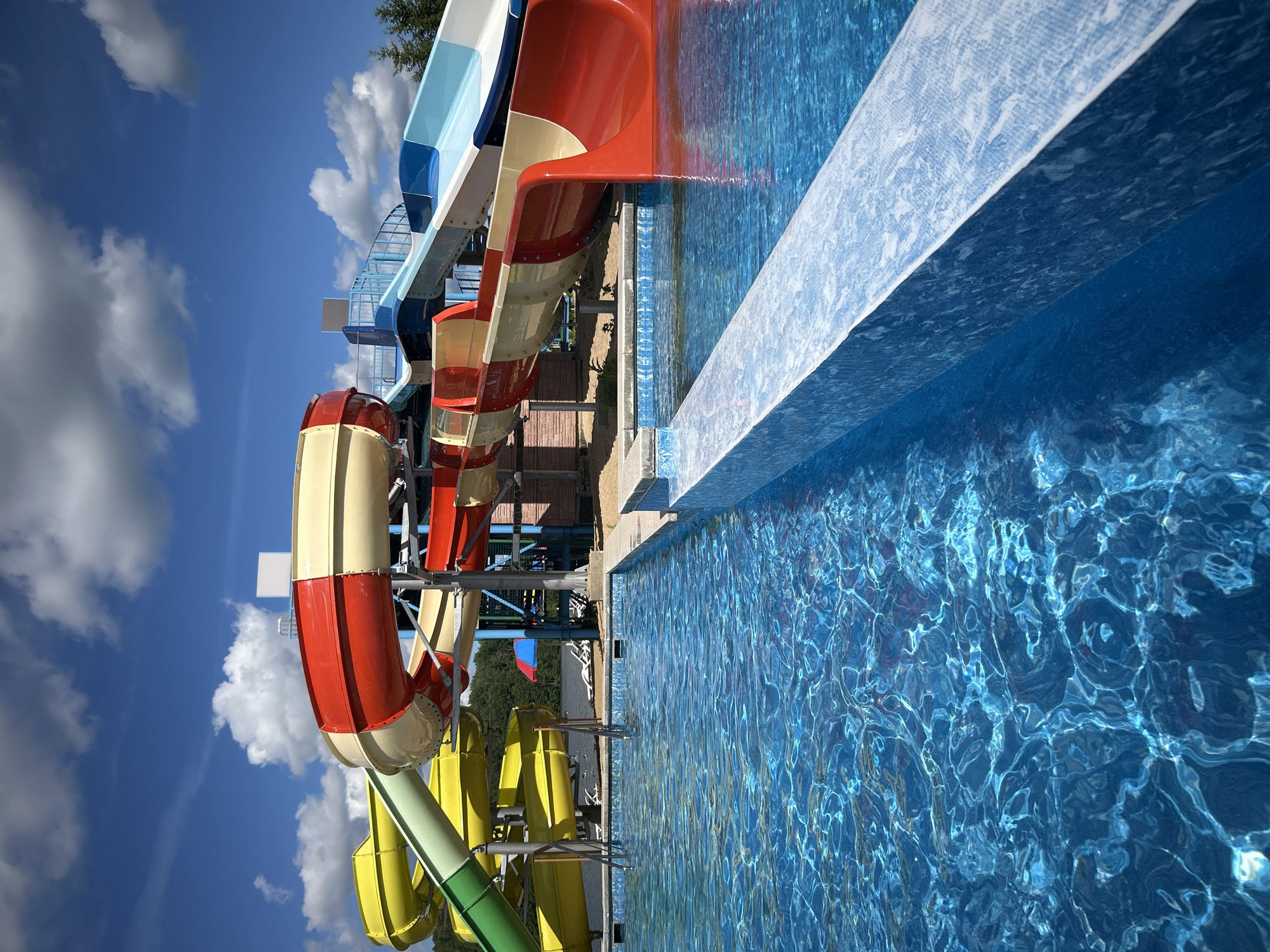 The image depicts a water park on a sunny day, with bright blue skies and white, fluffy clouds overhead. In the foreground, a section of a shimmering blue swimming pool is visible, with sunlight creating intricate patterns on its surface. A tiled edge of the pool forms a clean line, leading the eye towards the main attraction: a complex of colorful water slides.
The slides are a dominant feature, showcasing vibrant hues of red, yellow, green, and blue. Some are enclosed tubes, while others are open flumes. They are supported by a metal framework, suggesting a multi-level structure. In the background, beyond the slides, a line of trees can be seen, hinting at a natural setting. A few lounge chairs and a small red and blue umbrella are scattered around the base of the slides, implying a place for relaxation.
There are no people visible in the image. The scene is static and focuses on the architecture and environment of the water park. The overall impression is one of a recreational facility, ready for visitors on a warm day. The location is identified as the city of Юбилейный, Russia.