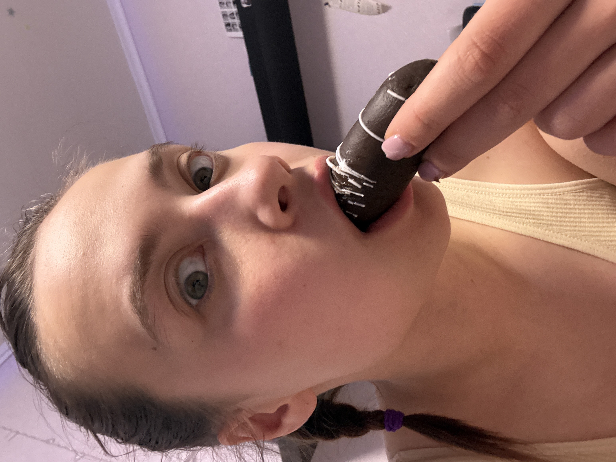 The image captures a close-up, angled view of a young female in an indoor setting, facing the viewer directly. She is in the process of eating a dark brown, elongated pastry, possibly a chocolate eclair or similar cake, which is decorated with thin white icing drizzles. Her right hand, featuring light-colored nail polish, holds the pastry to her mouth, which is open around its end.
The female subject has fair skin, wide-open, light-colored eyes (appearing greenish-grey), and dark hair. A braid secured with a purple elastic band is visible near her shoulder. She is wearing a light-colored, possibly cream or yellow, ribbed top. Her expression, characterized by her wide eyes, appears to be one of surprise, playfulness, or an exaggerated reaction to consuming the pastry.
The background is indistinct and out of focus, suggesting an interior room with softly lit, light-colored walls, possibly pale purple or white. There are no specific objects or details in the background that would provide further location cues within Прикамский, Russia. No visible text is present in the image. The lighting appears to be ambient indoor light.
