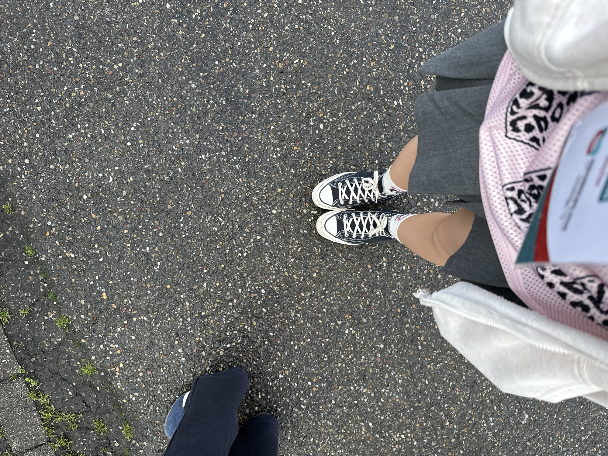 A first-person overhead shot shows a person wearing black Converse sneakers, grey shorts, a pink mesh top with a black and white animal print design, and white socks, standing on a dark grey asphalt surface. A second person wearing dark blue pants and matching shoes is partially visible in the lower left corner of the frame. The asphalt shows a mix of small, light-colored pebbles. Some small patches of grass are visible on the edge of the paved surface. The location is in Strasbourg, France.