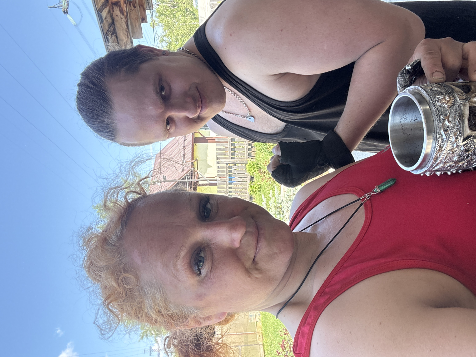 A medium-shot, selfie-style photograph depicts two individuals standing outdoors on a sunny day, likely in a backyard or residential area. The woman in the foreground is positioned on the left, facing the camera with a slight smile. She has reddish-blonde curly hair pulled back, and is wearing a red tank top with a black cord necklace adorned with a green pendant. There is a visible bruise on her left cheek. To her right and slightly behind her stands a man wearing a black tank top and a silver chain necklace. He has dark hair pulled back and is smirking at the camera. His right hand is clenched in a fist, wearing a black glove or wrap. He is holding a decorative metal mug with an ornate handle in his left hand. The background suggests a rural or suburban setting, with a wooden fence, greenery, and residential structures visible. The sky is clear and blue with some wispy clouds. The overall impression is informal, possibly taken during a casual outdoor gathering or activity. This location is identified as being in the city of Chaikovsky, Russia.