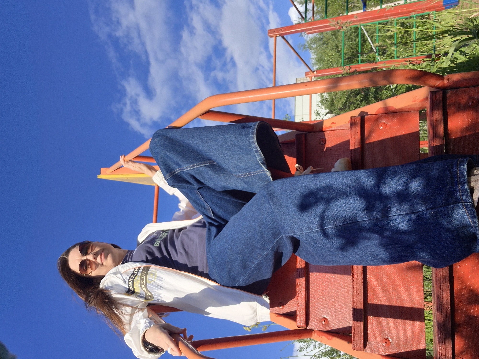 The image displays a young woman reclining on a red and orange playground structure on a bright, sunny day, likely in Chaikovsky, Russia. She is positioned with her torso resting on a lower part of the structure, and her legs extended upwards along what appears to be a slide or climbing frame.
The woman has long, dark hair and is wearing brown-tinted sunglasses. Her clothing includes a dark blue t-shirt with faint white text, and oversized, wide-leg blue denim jeans. A white long-sleeved top or jacket, featuring stylized black and white text (parts of which resemble "SUISWXME"), is draped around her. Her hands are gently gripping the orange bars of the playground equipment.
The scene is outdoors under a clear, vibrant blue sky dotted with scattered white clouds. Lush green foliage, including trees and grass, is visible in the background, suggesting a park or a public recreational area. The strong sunlight indicates it is daytime, possibly mid-morning or afternoon. The overall impression is one of leisure and relaxation in an outdoor setting.