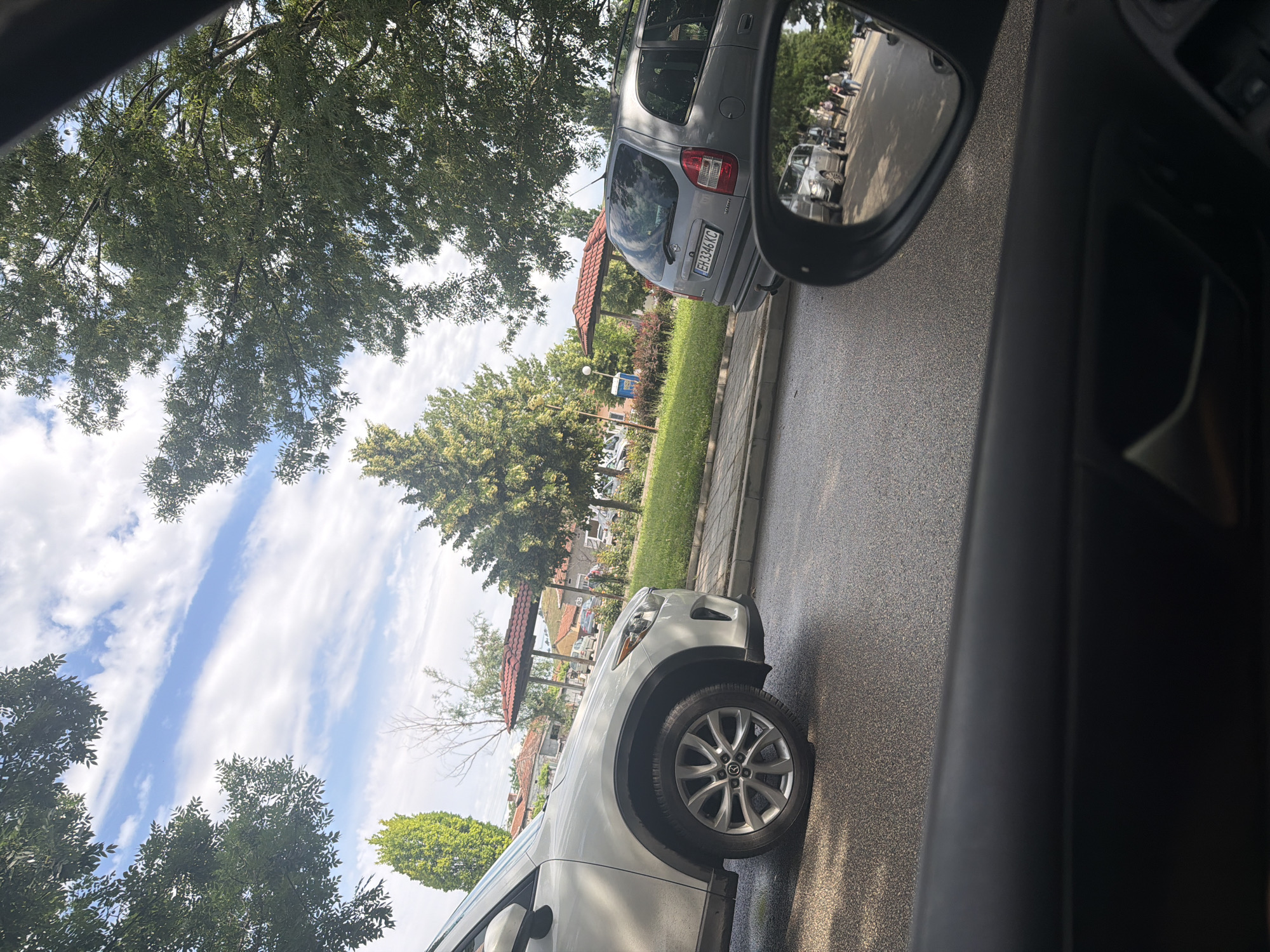 This image, captured from within a vehicle and originally rotated, presents a daytime street scene in Iskar, Bulgaria. The perspective looks out over an asphalt road and sidewalk, with a blend of natural elements and parked cars.
In the foreground, the dark interior trim of the observer's vehicle is visible on the far right. The front section, wheel, and tire of a white SUV occupy the lower-left portion of the frame, indicating the car from which the photo was taken. The road surface appears dark and possibly damp or shiny.
Further down the street, a gray Opel Zafira station wagon is parked on the right, facing away from the camera. Its Bulgarian license plate, "EH 3346 KC," is clearly visible. Behind this car, a small red-roofed structure, possibly a gazebo or covered seating area, stands within a patch of green grass. A street lamp is also visible nearby.
The vehicle's side mirror is prominent on the right, reflecting a broader view of the street behind. In the reflection, a group of people are gathered on a sidewalk or plaza area, along with additional parked cars, suggesting a social interaction or waiting scenario.
The sky above is bright blue with distinctive white, streaky clouds, indicative of fair weather. Numerous trees with dense green foliage are prominent throughout the image, framing the sky and lining the street. The sunlight suggests it is mid-day or early afternoon. The scene depicts a typical, quiet street environment.