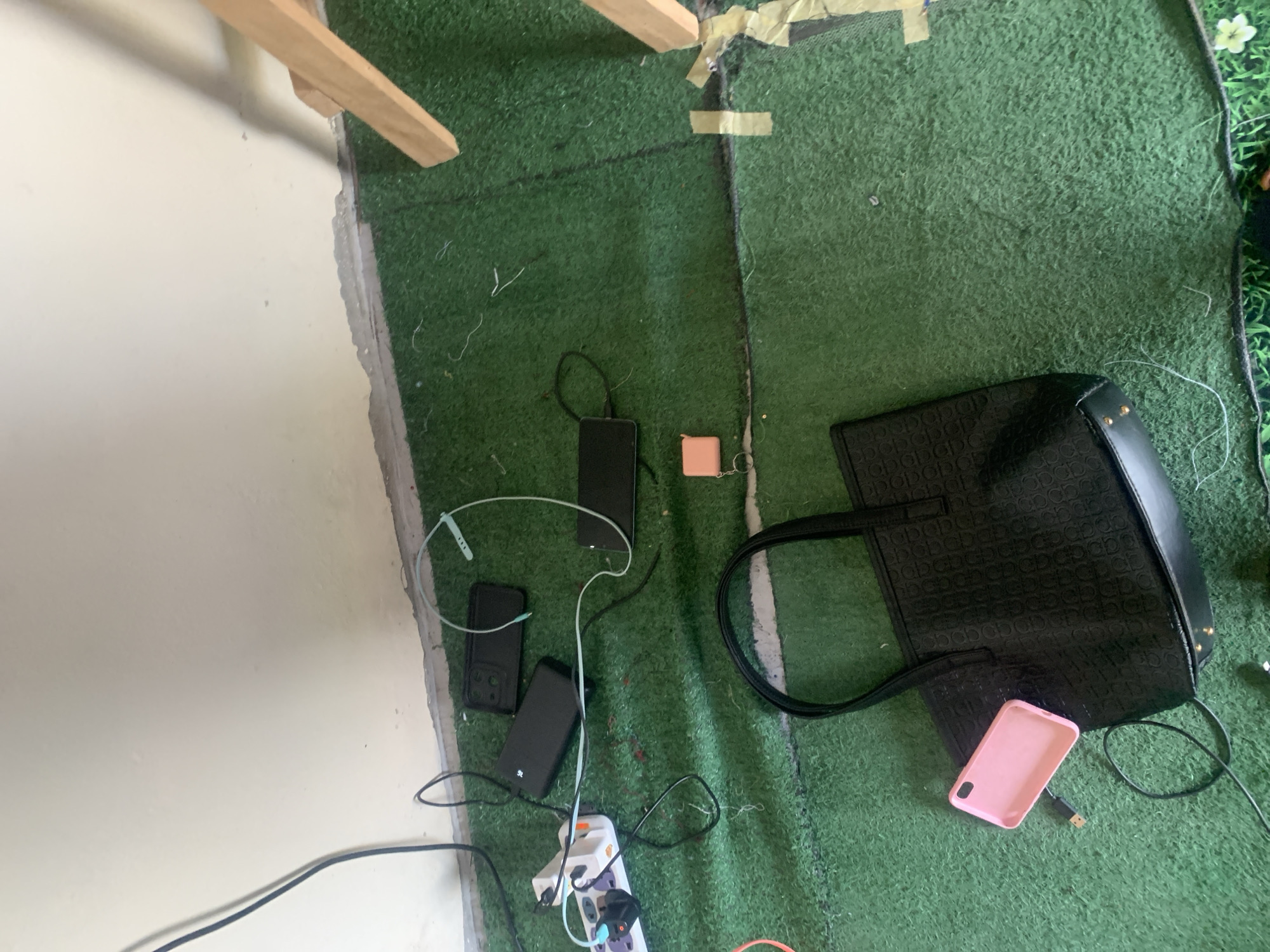 The image depicts a floor covered with artificial turf, appearing to be an indoor or sheltered outdoor space. Several electronic devices and accessories are scattered on the turf. A white power strip with multiple outlets is plugged in, with various black charging cables connected to it and extending across the surface. Among the items are at least three black rectangular objects, likely smartphones or power banks, with some cables attached. A light blue earbud wire is also visible. In the foreground, a black tote bag with a textured pattern rests on the turf. A bright pink smartphone case lies next to the tote bag, with a black USB cable attached. A small, peach-colored square object with a keychain is also present. The scene is lit, and the overall impression is one of disarray or a workspace with many active charging devices. No people are visible in the image. The location is described as Jauro Yinu, Nigeria, though this context is external to the visual information provided in the image itself.
