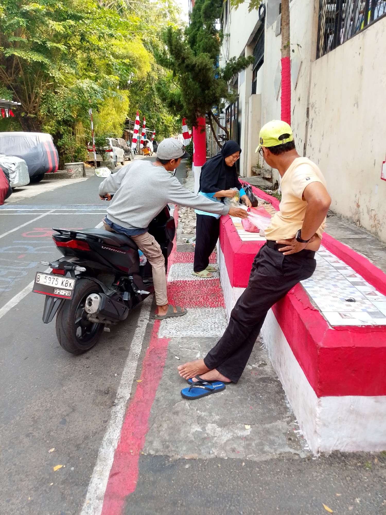 In East Jakarta, Indonesia, on a street with red and white painted markings, a man wearing a grey sweatshirt, light brown pants, and grey sandals is sitting on a black motorcycle. He is interacting with two other individuals. To his left, a woman in a black hijab and blue top is holding plastic bags, appearing to hand something to the man on the motorcycle. To his right, a man wearing a yellow baseball cap, a light yellow t-shirt, black pants, and blue flip-flops is sitting on a red and white tiled ledge, his left hand on his hip. The environment includes trees, a building with a white wall, and parked vehicles in the background.